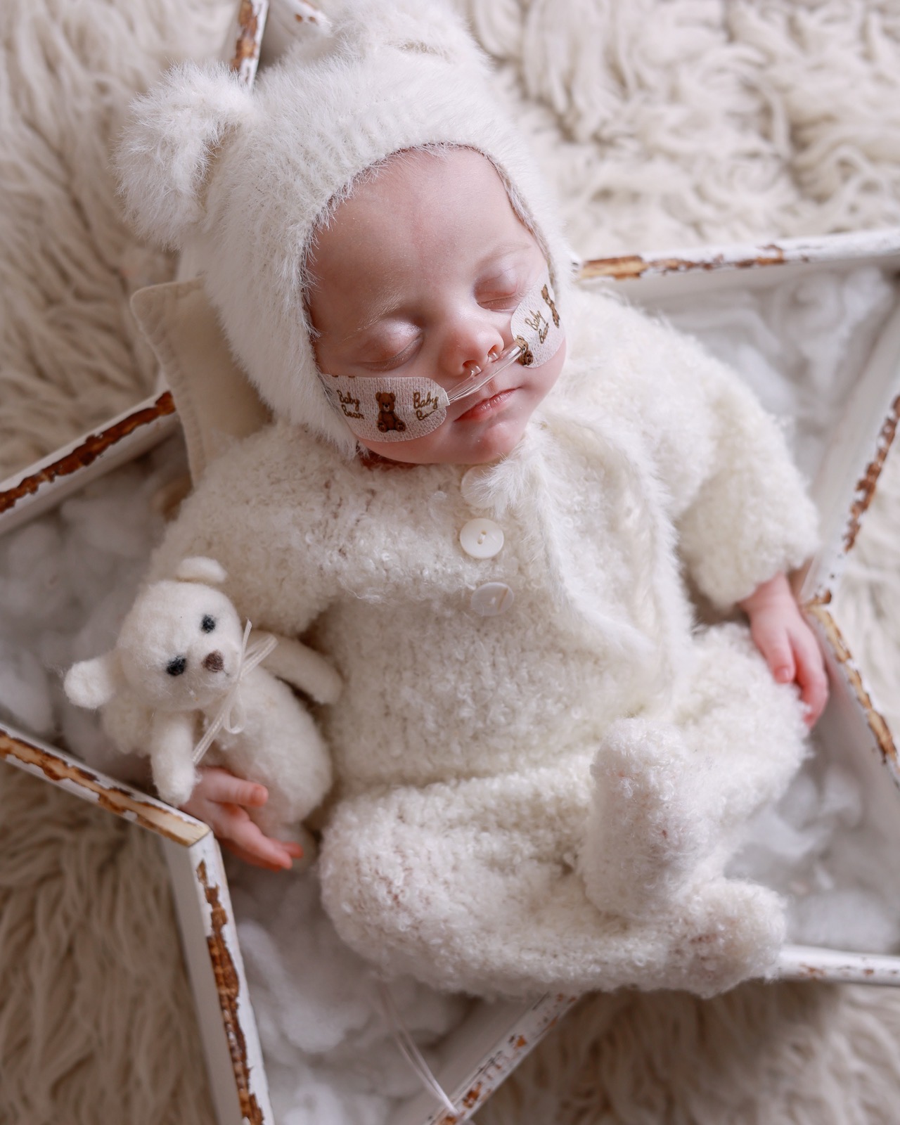Sleeping newborn boy in a cream knitted bear-eared bonnet with a nasal cannula, cuddling a small knitted teddy bear on a wooden star-shaped prop