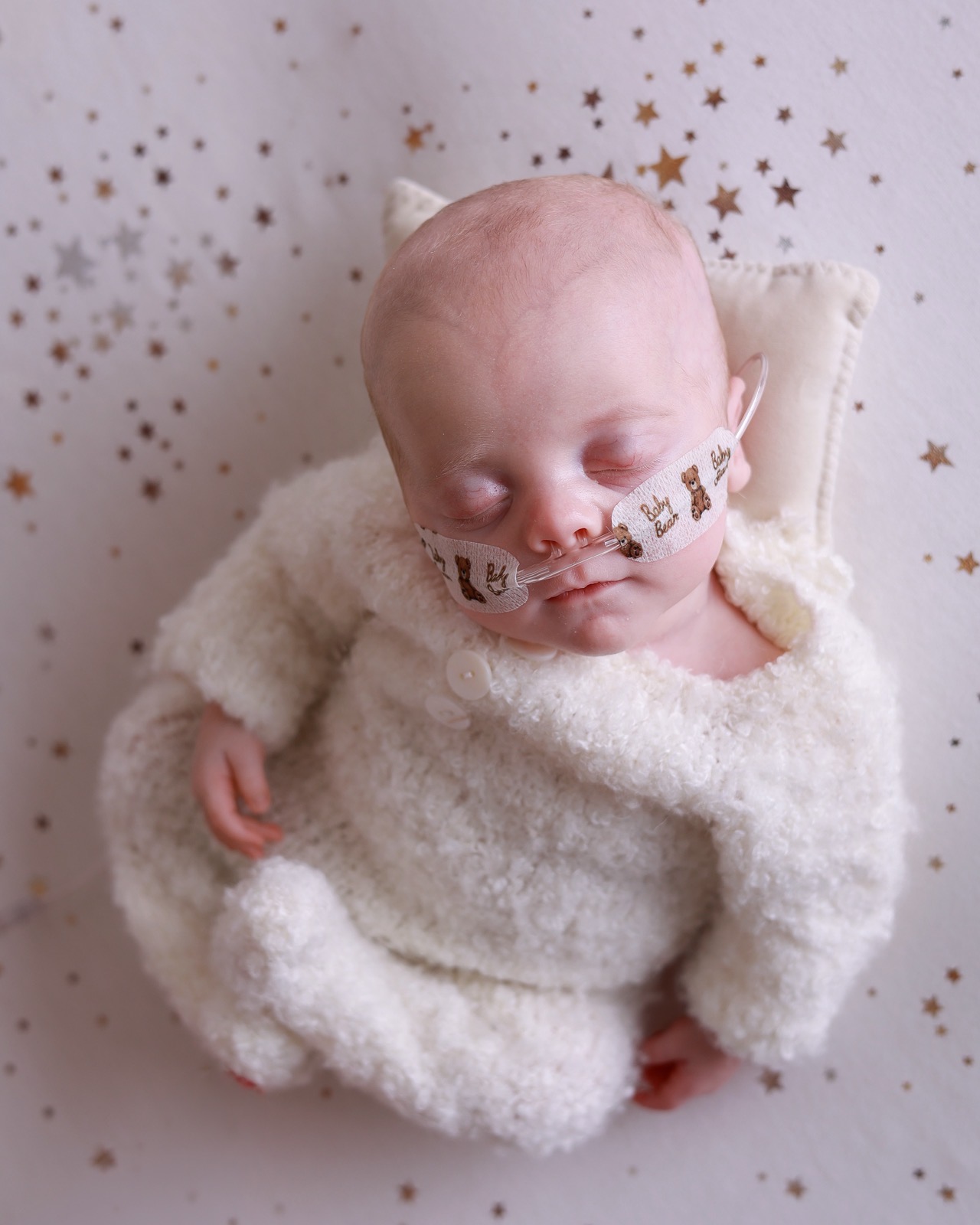 Sleeping newborn boy lying on his back in a cream knitted bear-eared bonnet with a nasal cannula, star-speckled backdrop