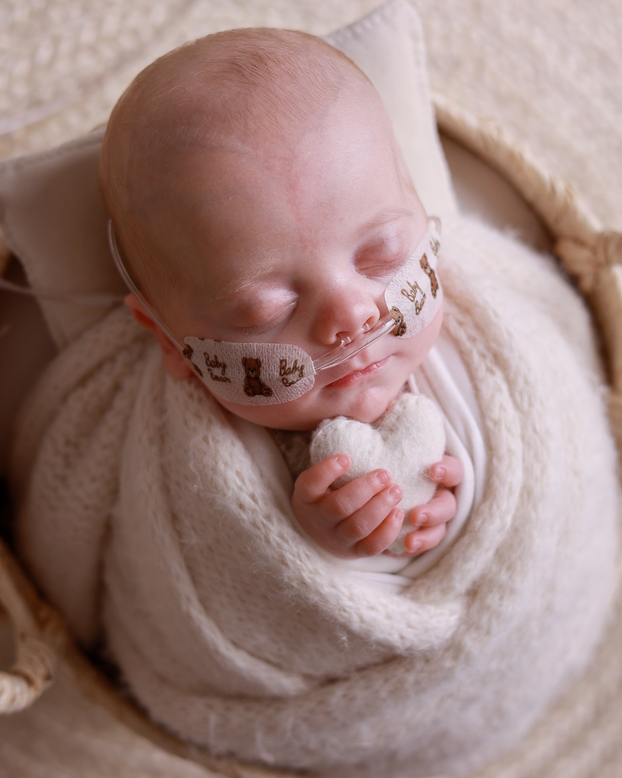 Sleeping newborn boy swaddled in a cream wrap in a woven basket, holding a small felt heart, wearing a nasal cannula