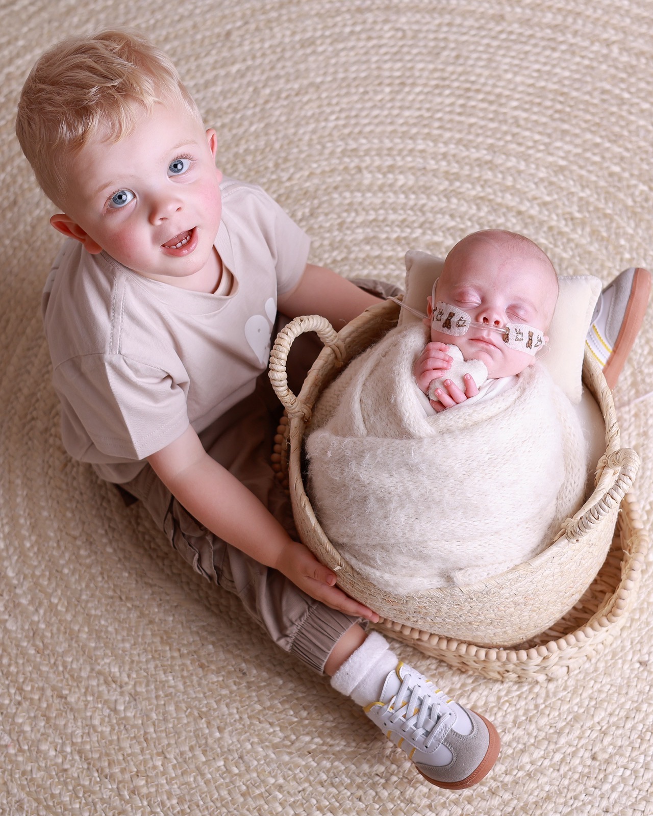 Older brother kneeling proudly beside a woven basket holding his sleeping newborn sibling, who wears a nasal cannula