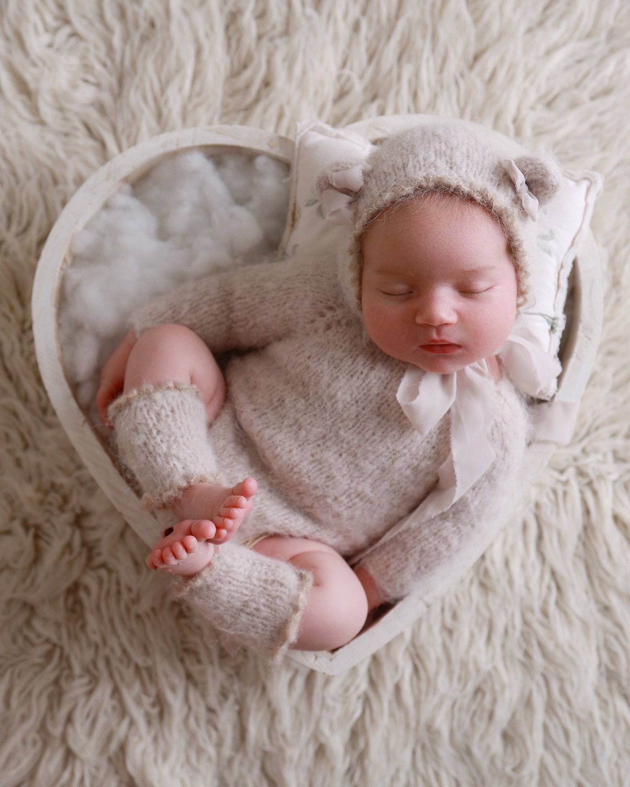 Sleeping newborn girl in a taupe knitted bear-eared bonnet, lying in a heart-shaped wooden prop