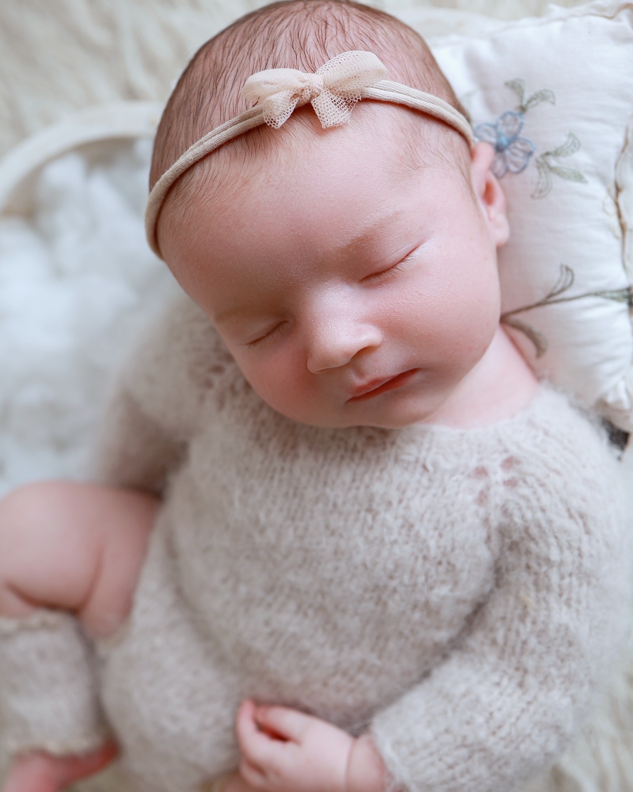 Sleeping newborn girl in a taupe knitted outfit and bow headband, resting on an embroidered floral cushion