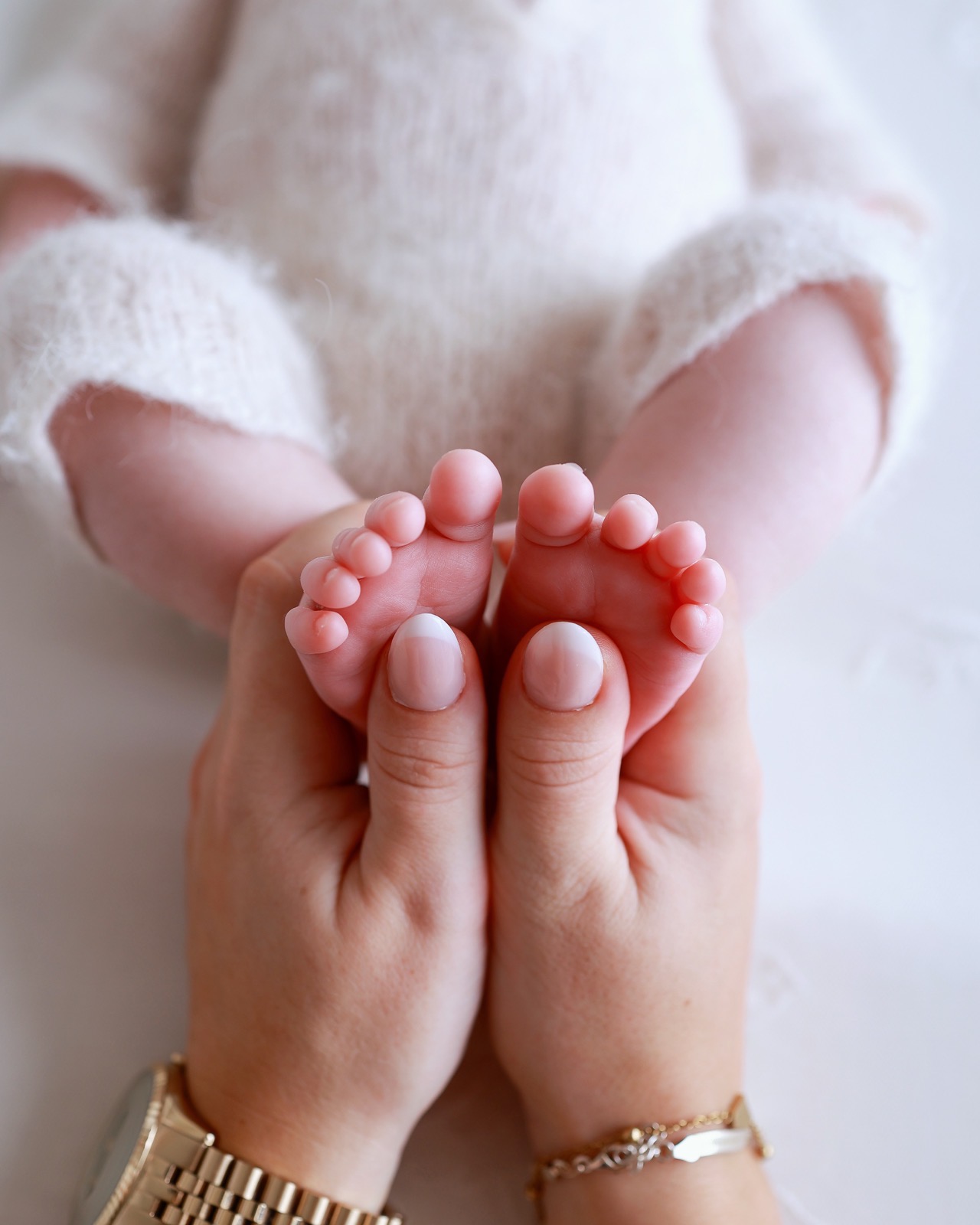 Close-up of a parent's hands cradling their newborn's two feet together, wearing a gold watch and bracelet