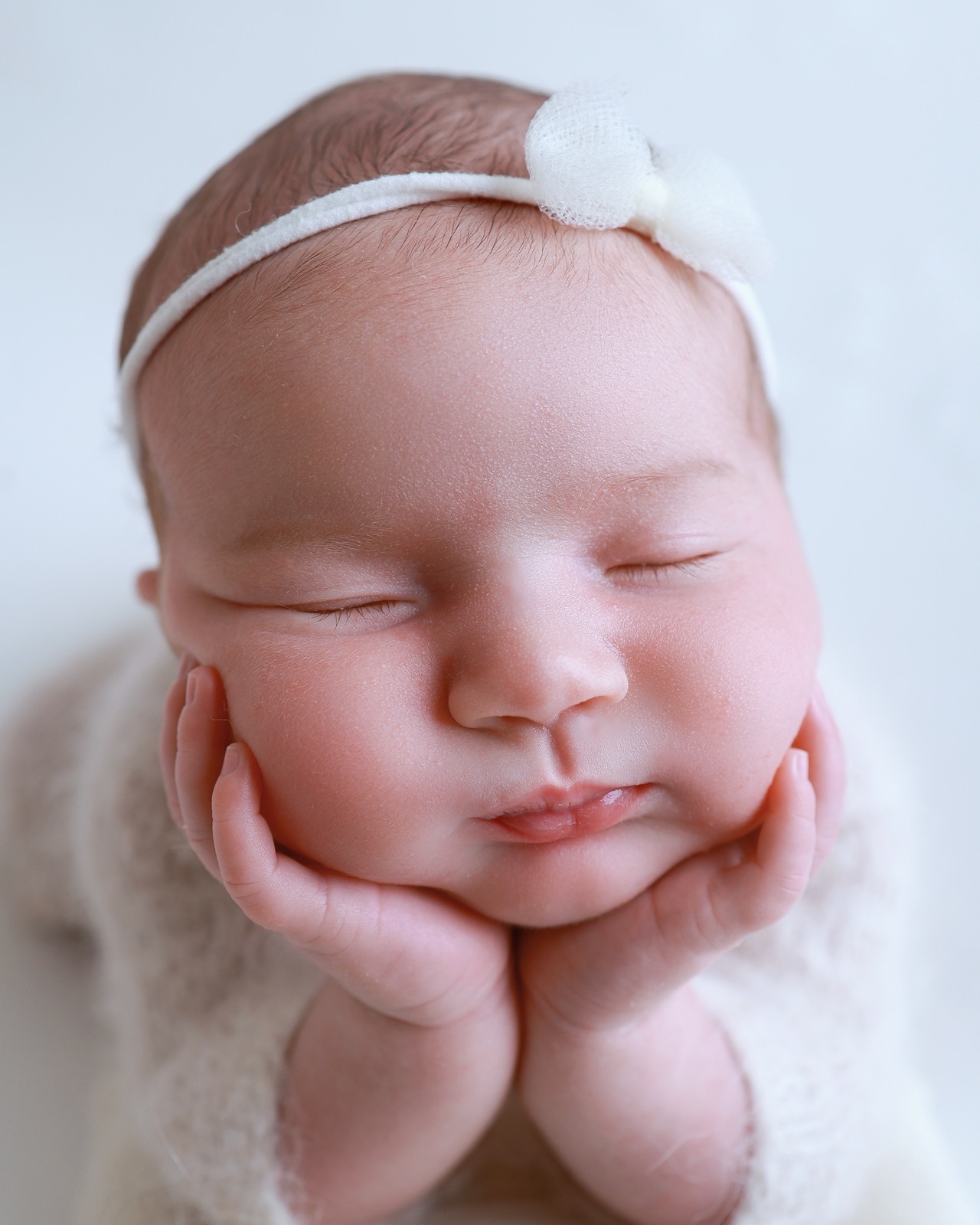 Close-up of a sleeping newborn resting her chin on her hands, white bow headband, cream knitted outfit