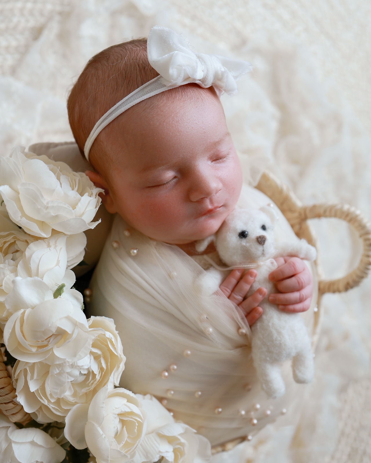 Sleeping newborn swaddled in a pearl-beaded wrap, holding a small knitted teddy bear, surrounded by white roses