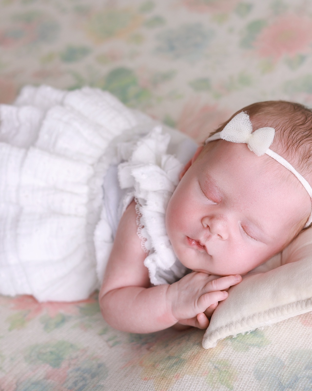 Sleeping newborn in a white ruffled outfit and bow headband, lying on a soft floral-print blanket