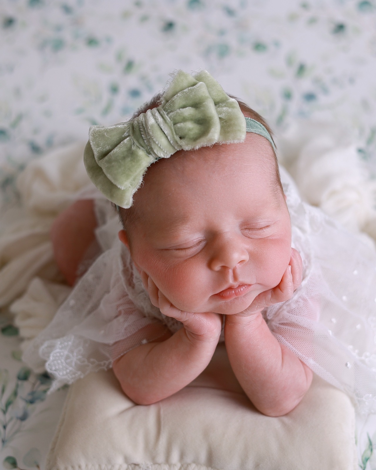 Sleeping newborn resting her chin on her hands, sage green velvet bow headband, white lace outfit, floral backdrop