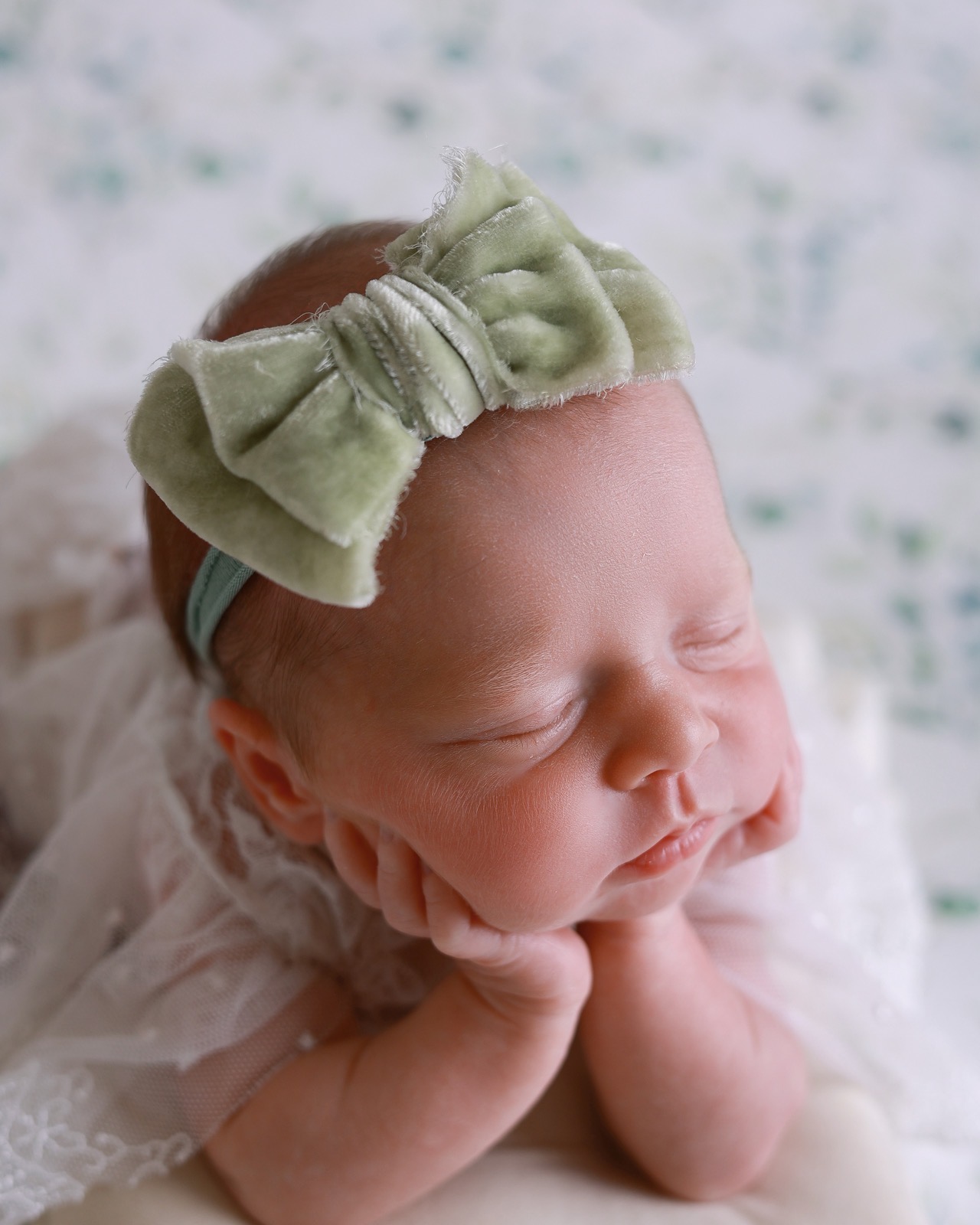 Sleeping newborn with a sage green fabric flower headband, arms resting either side, floral eucalyptus-print backdrop