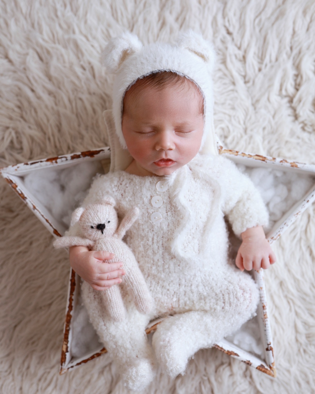 Sleeping newborn boy in a cream knitted bear-eared bonnet lying on a wooden star prop, holding a small knitted teddy bear
