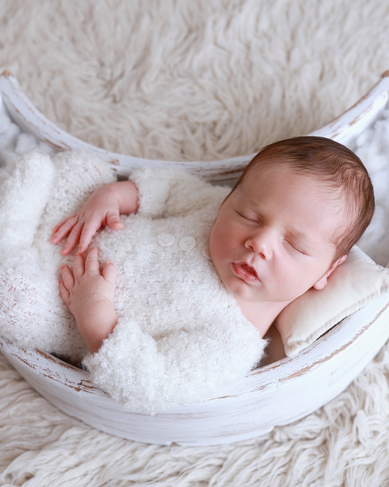 Sleeping newborn boy in a cream knitted romper lying in a white wooden crescent-moon prop