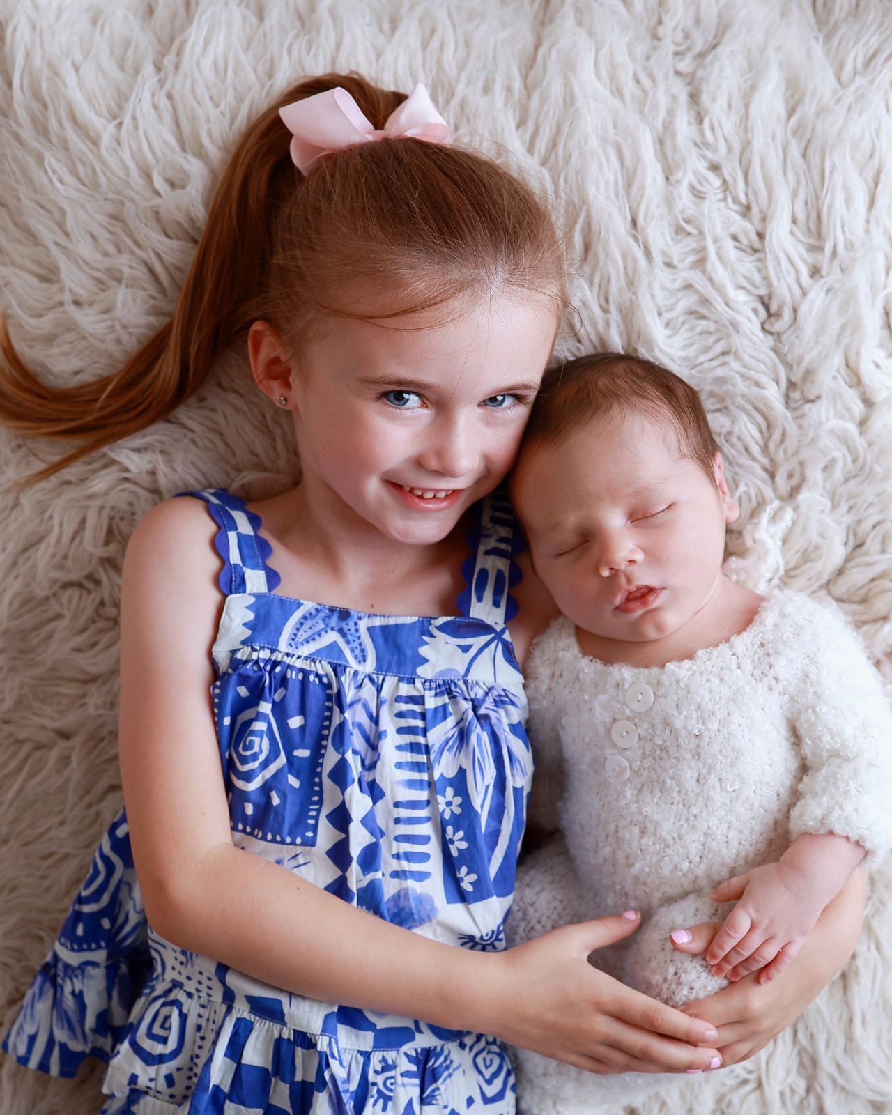 Older sister lying beside her sleeping newborn sibling, both in cream knitted outfits, on cream faux fur