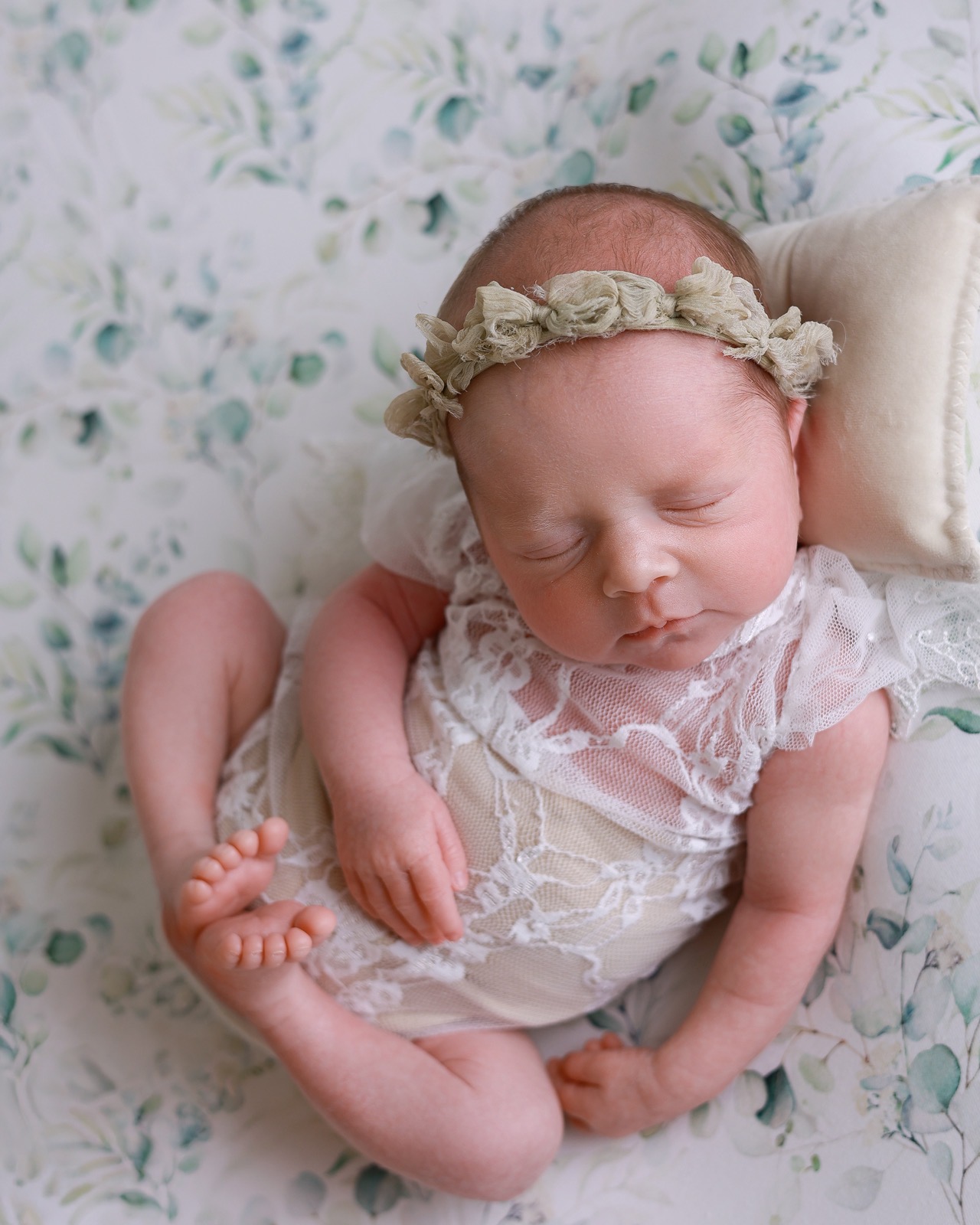 Sleeping newborn girl with a sage green fabric flower headband, cream lace romper, floral eucalyptus-print backdrop