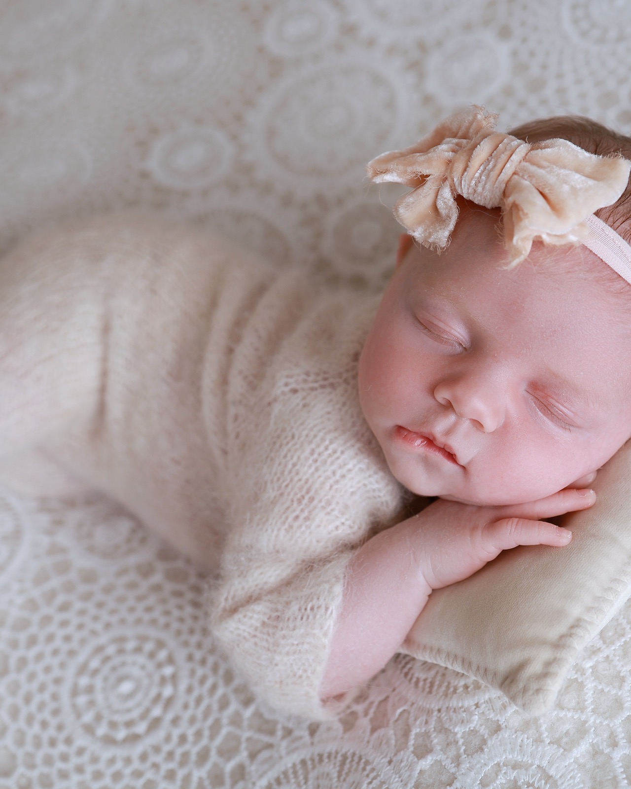 Sleeping newborn girl with a peach velvet bow headband, cream knitted outfit, resting on a lace blanket