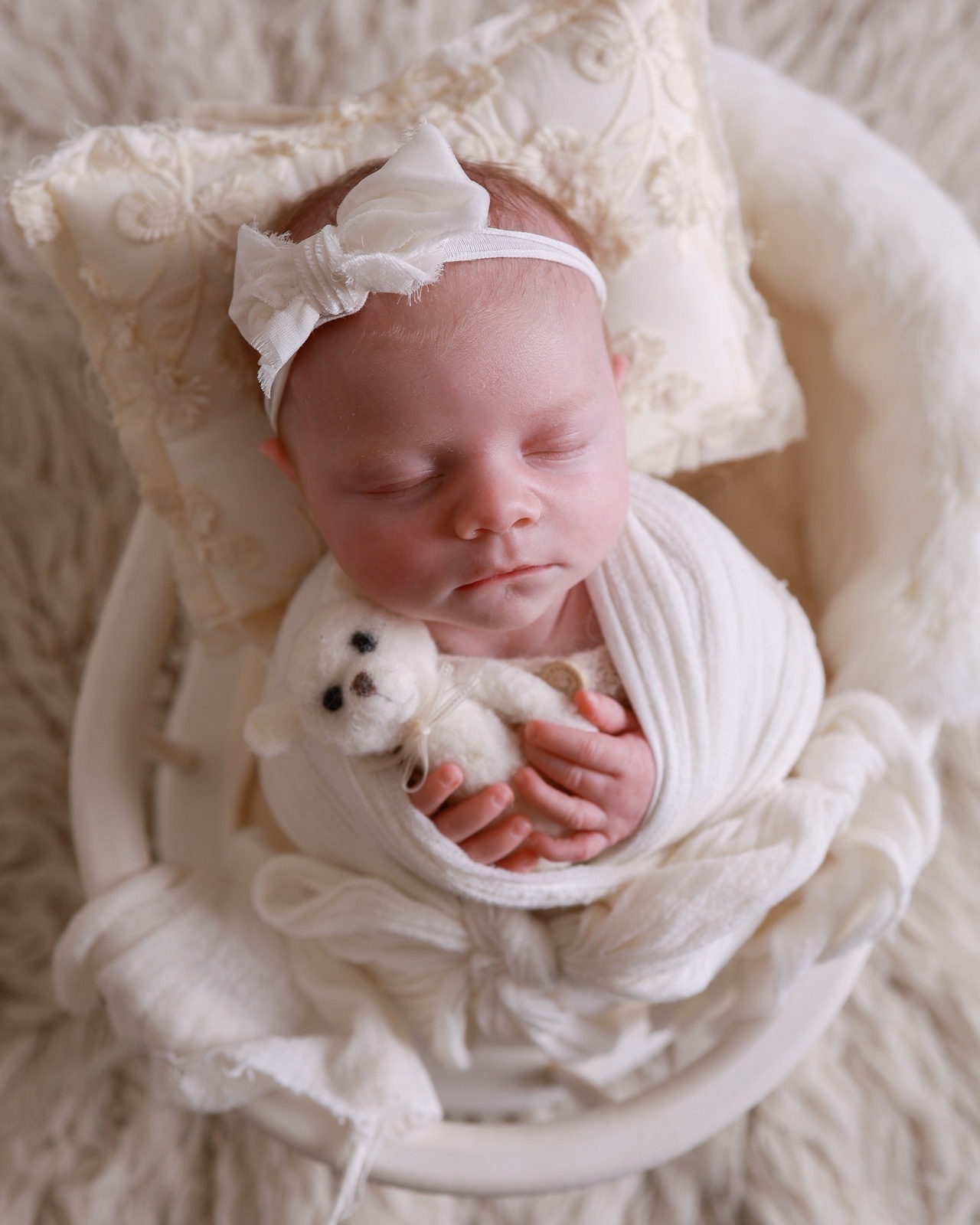 Sleeping newborn swaddled in a cream wrap, white bow headband, holding a small knitted teddy bear, in a round basket