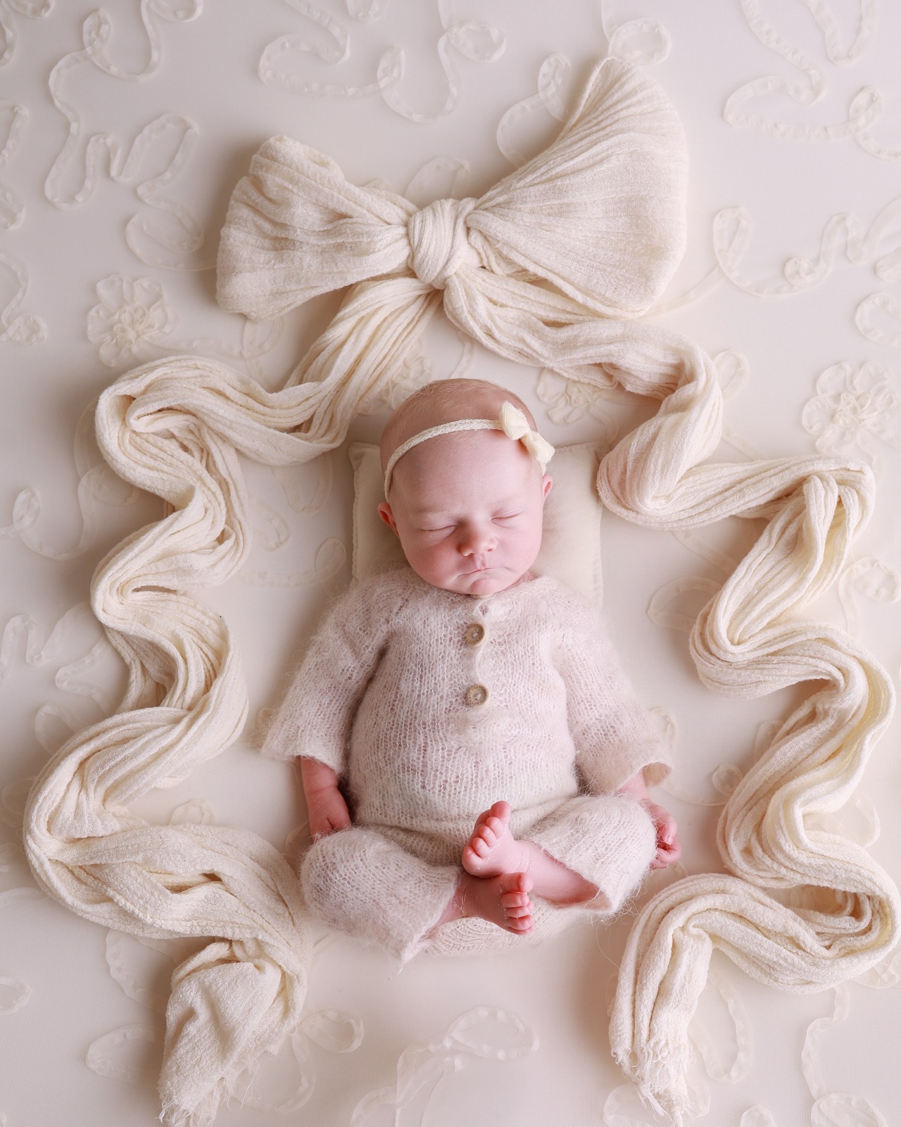 Sleeping newborn sitting cross-legged in a cream knitted outfit, framed by a decorative ribbon bow