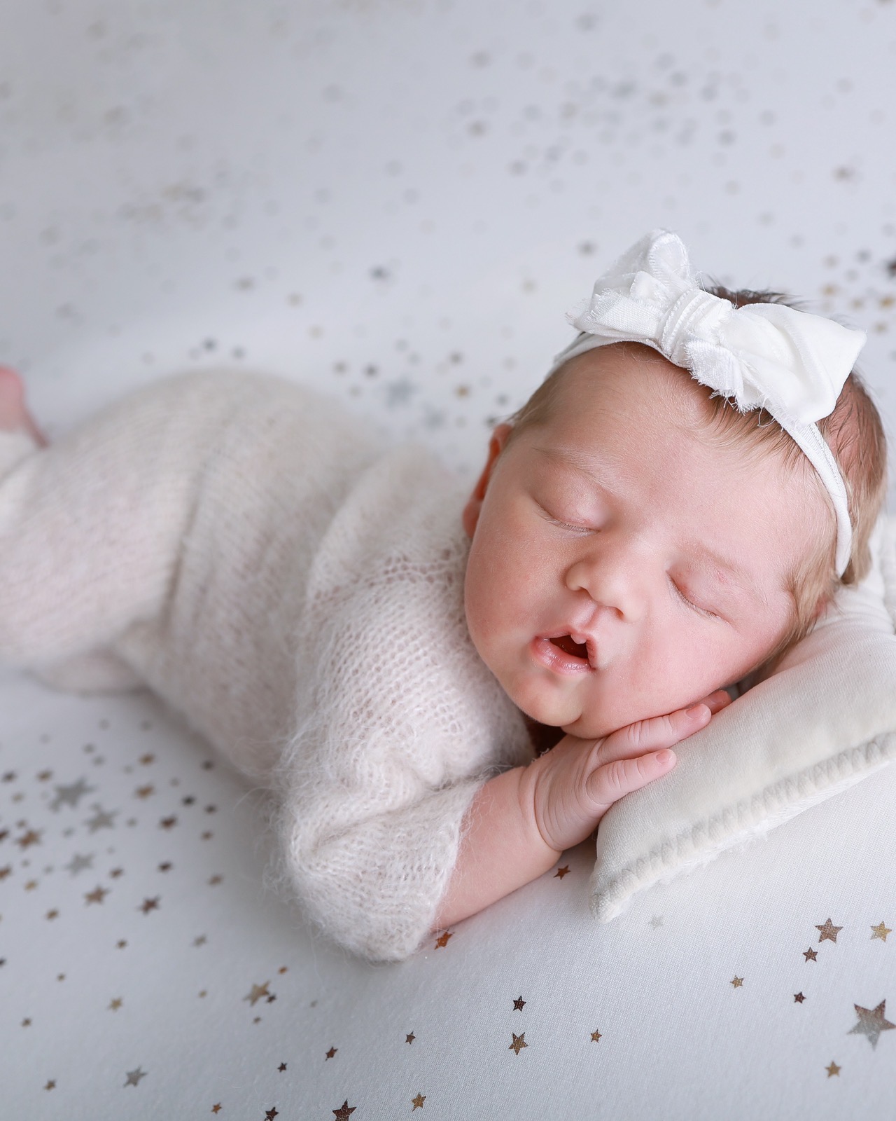 Sleeping newborn lying on her side, cream knitted outfit and bow headband, mouth slightly open, star-speckled backdrop