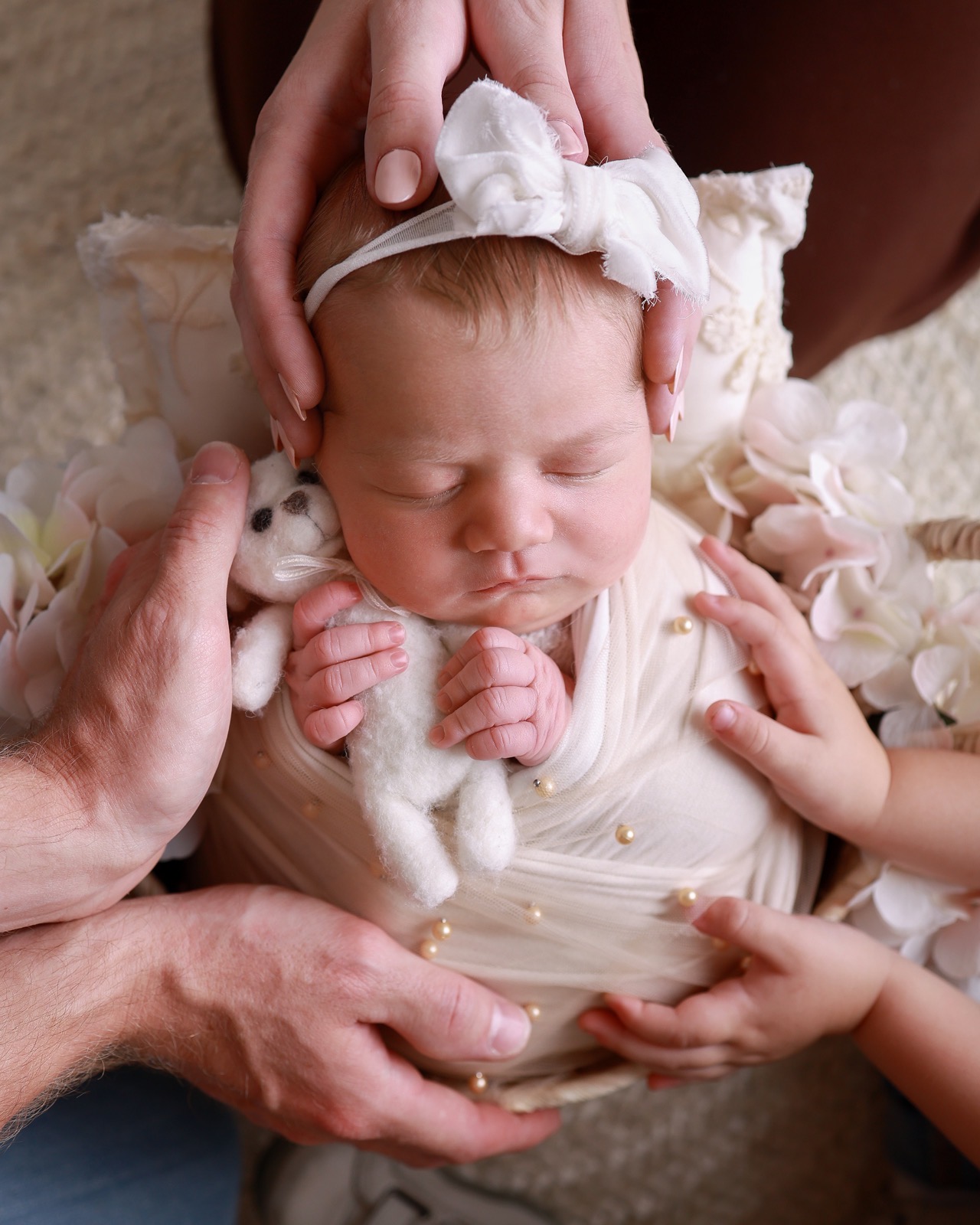 A family's hands all resting gently on a sleeping newborn wrapped in cream with pearls, small white flowers nearby
