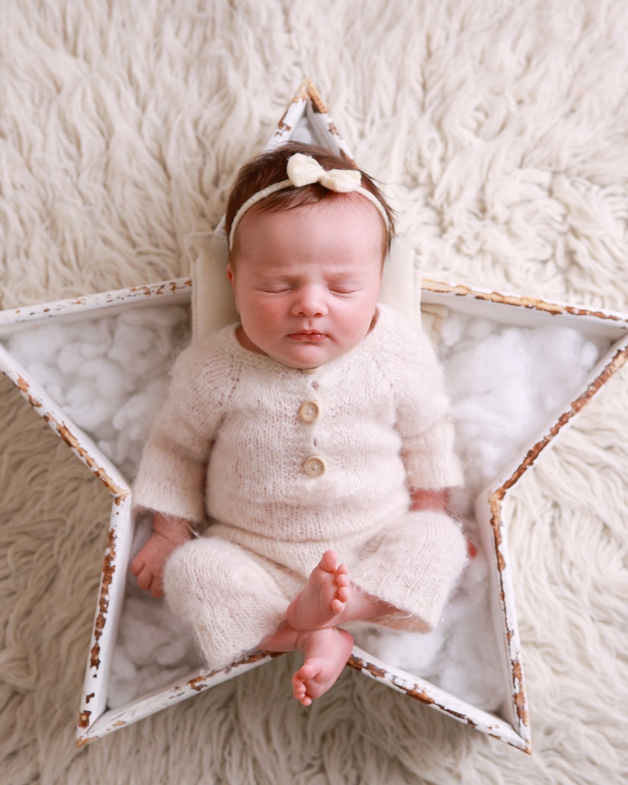 Sleeping newborn sitting cross-legged in a white star-shaped prop, cream knitted outfit and bow headband