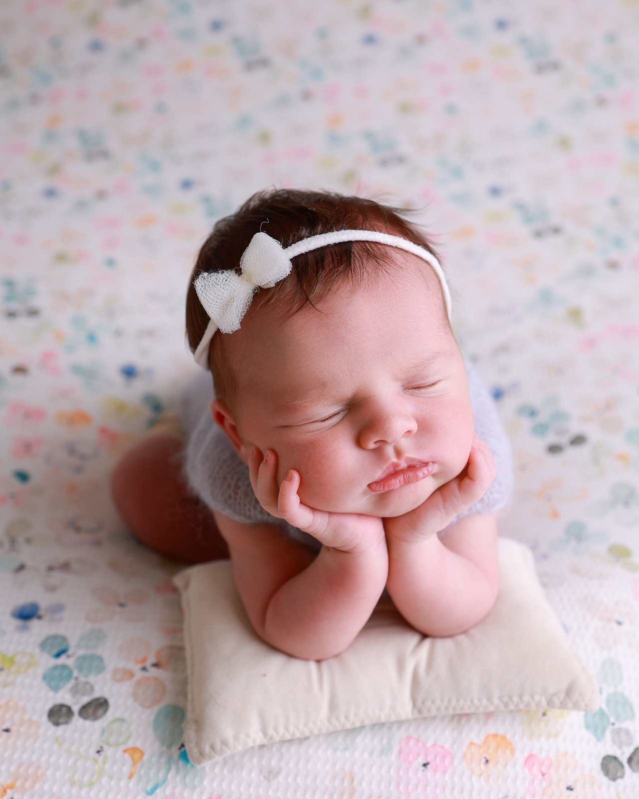 Close-up of a sleeping newborn resting her chin on her hands, lilac-grey knitted outfit, floral speckled backdrop