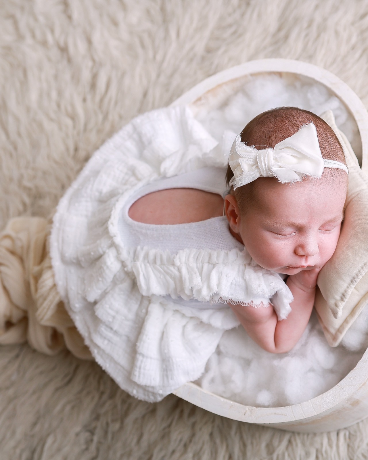 Sleeping newborn lying in a round wooden bowl, white ruffled outfit and bow headband, hand near her mouth