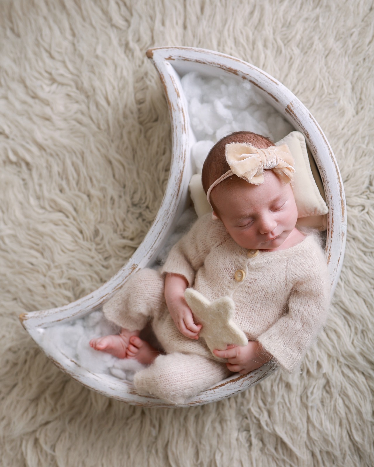 Sleeping newborn girl in a crescent-moon prop, oatmeal knitted outfit and velvet bow headband, holding a small felt star