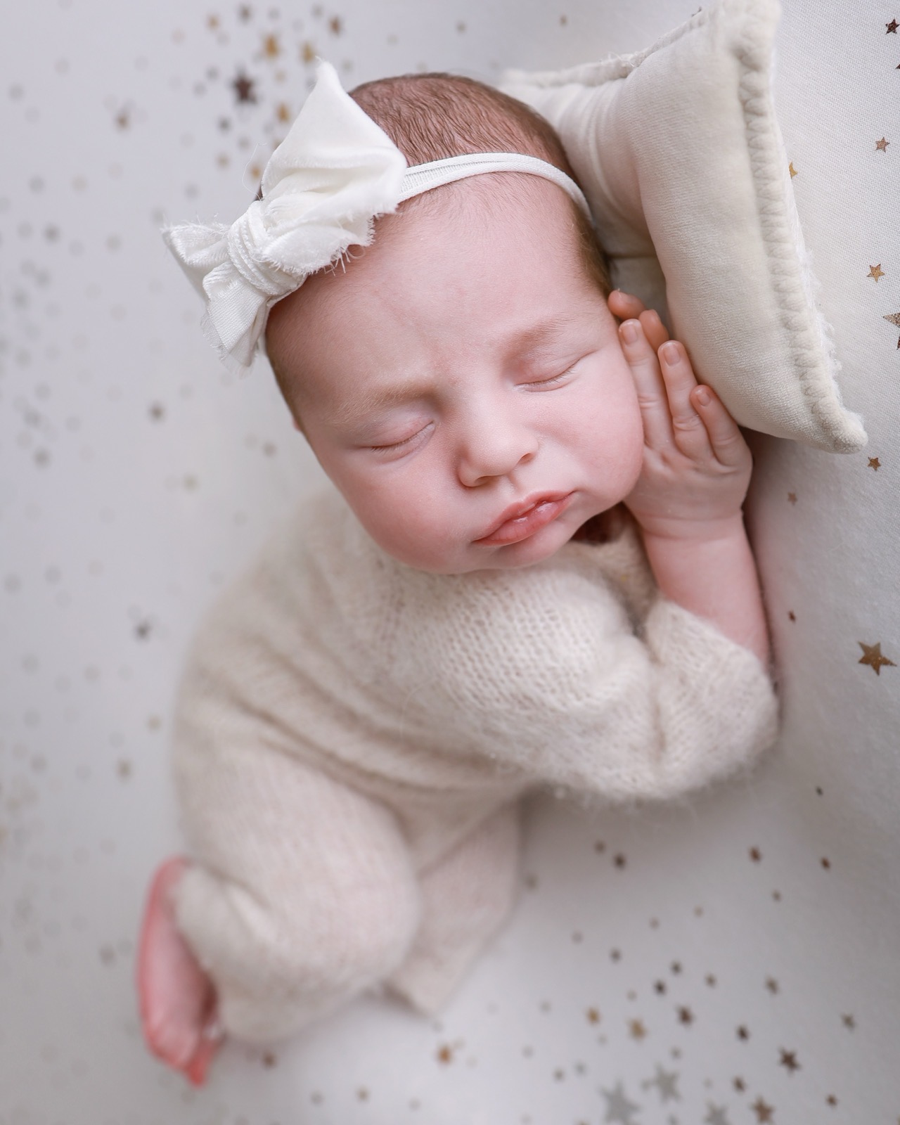 Sleeping newborn in side profile, hand near her cheek, cream bow headband, gold star-speckled backdrop