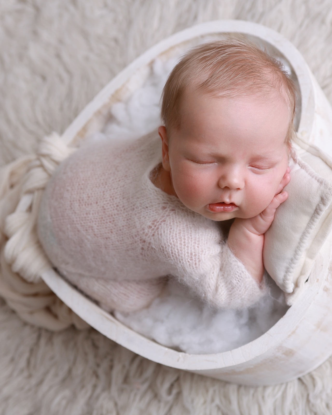 Sleeping newborn lying in an oval basket, pink knitted outfit, chin resting on her hands atop a cream cushion