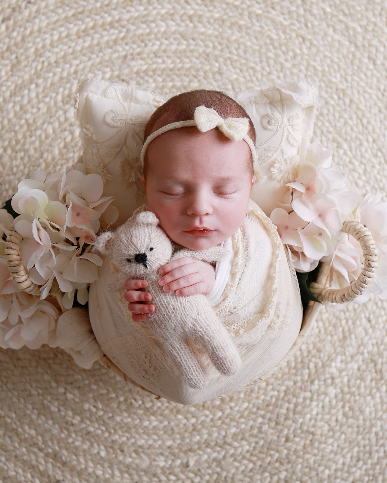 Sleeping newborn swaddled in cream lace, bow headband, holding a small knitted teddy bear, surrounded by pale pink hydrangeas