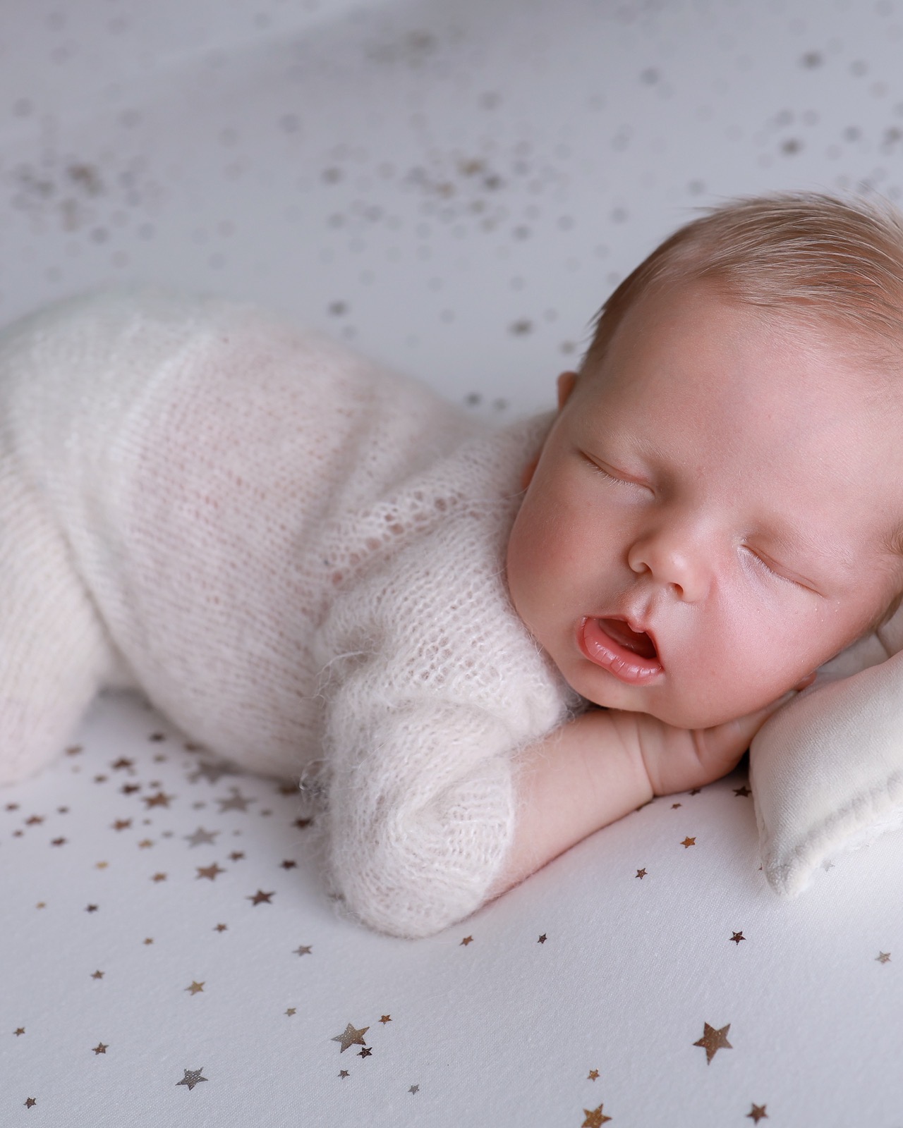 Sleeping newborn on her side, mouth slightly open, cream knitted outfit, gold star-speckled backdrop