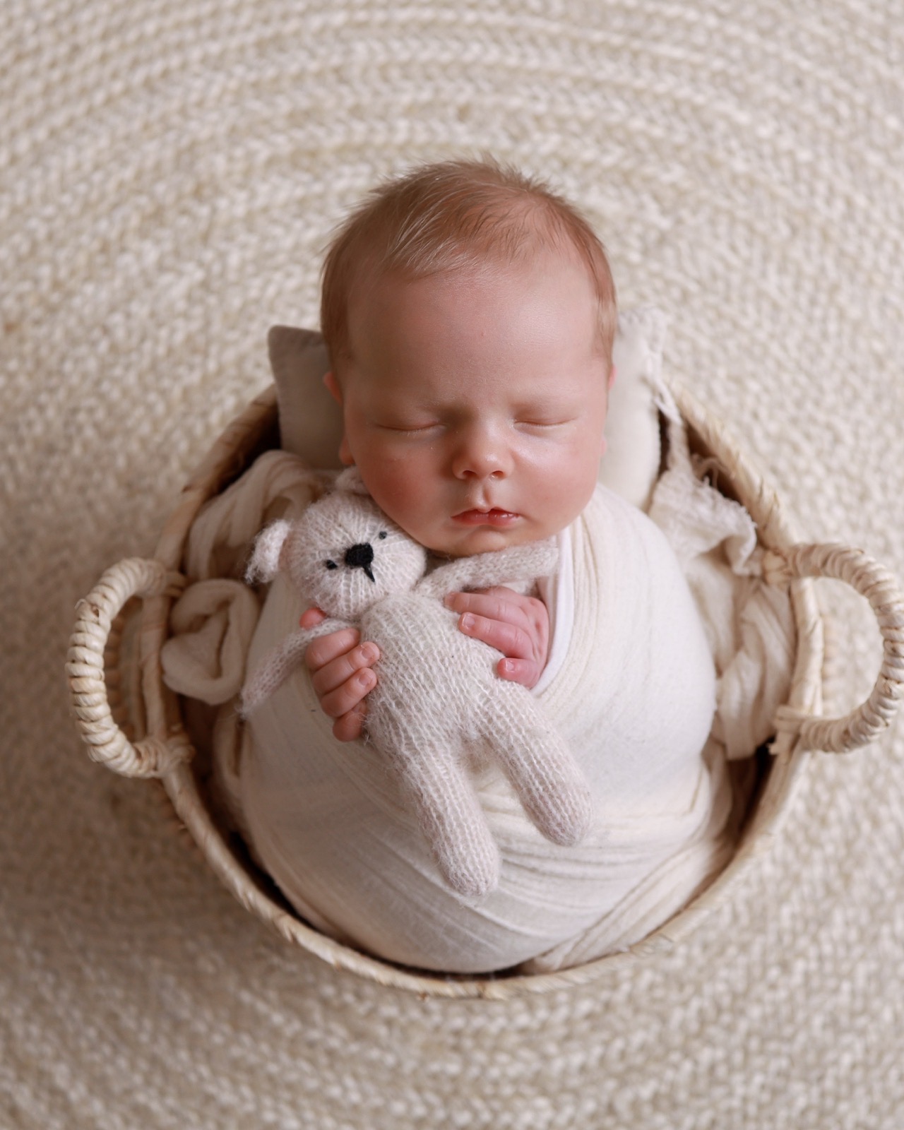 Sleeping newborn boy swaddled in a cream wrap in a woven basket, holding a small knitted teddy bear