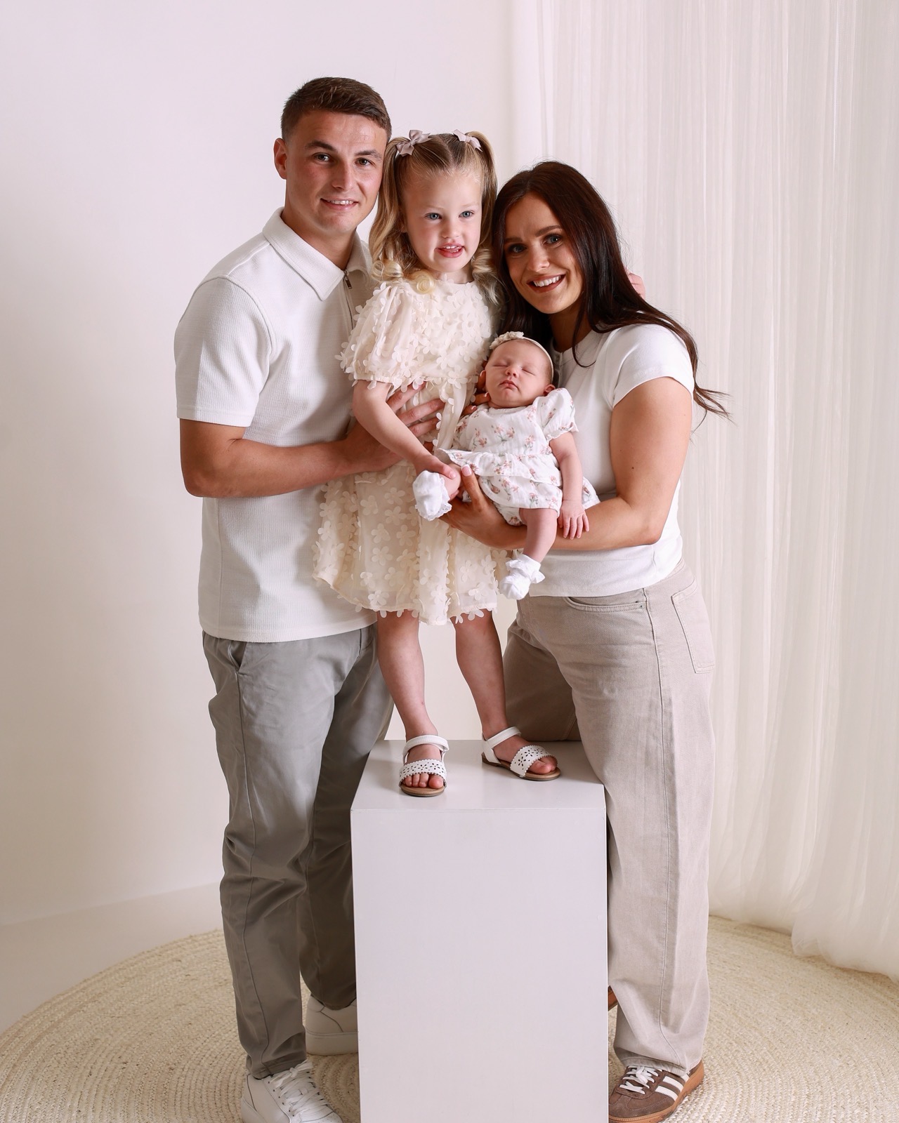 Family of four, older daughter in a cream floral dress standing on a white plinth, parents holding their sleeping newborn