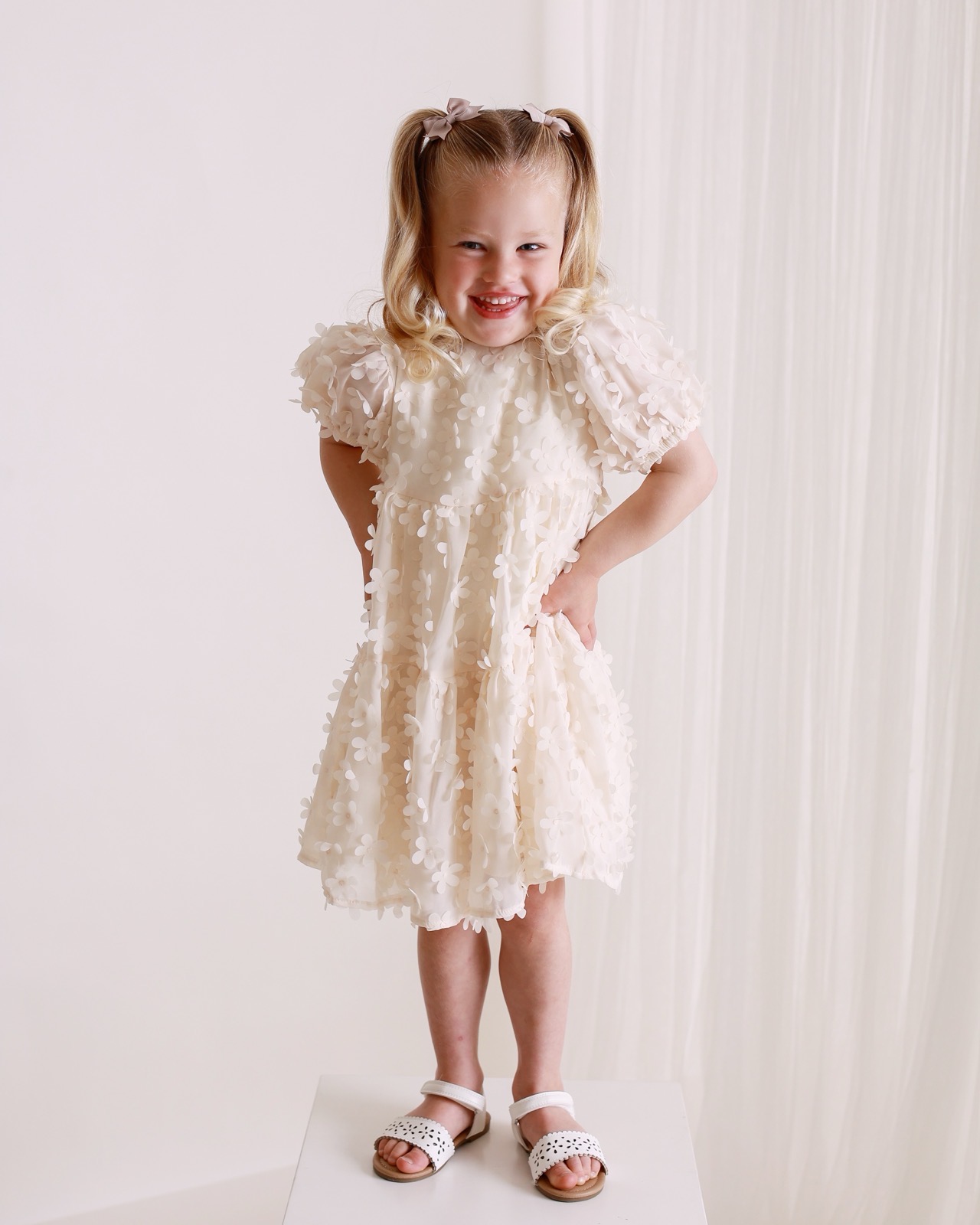 Young girl in a cream floral puff-sleeve dress and hair bows, standing on a white plinth, smiling