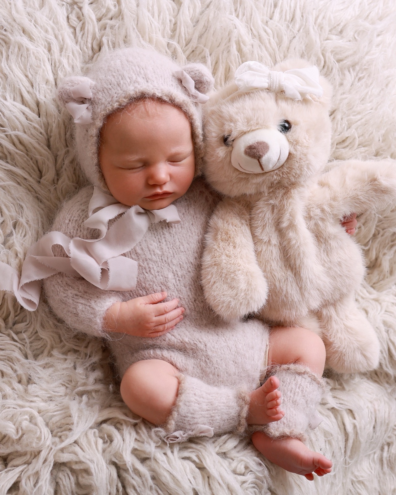 Sleeping newborn in a taupe knitted bear-eared bonnet, cuddled up to a cream teddy bear toy on faux fur