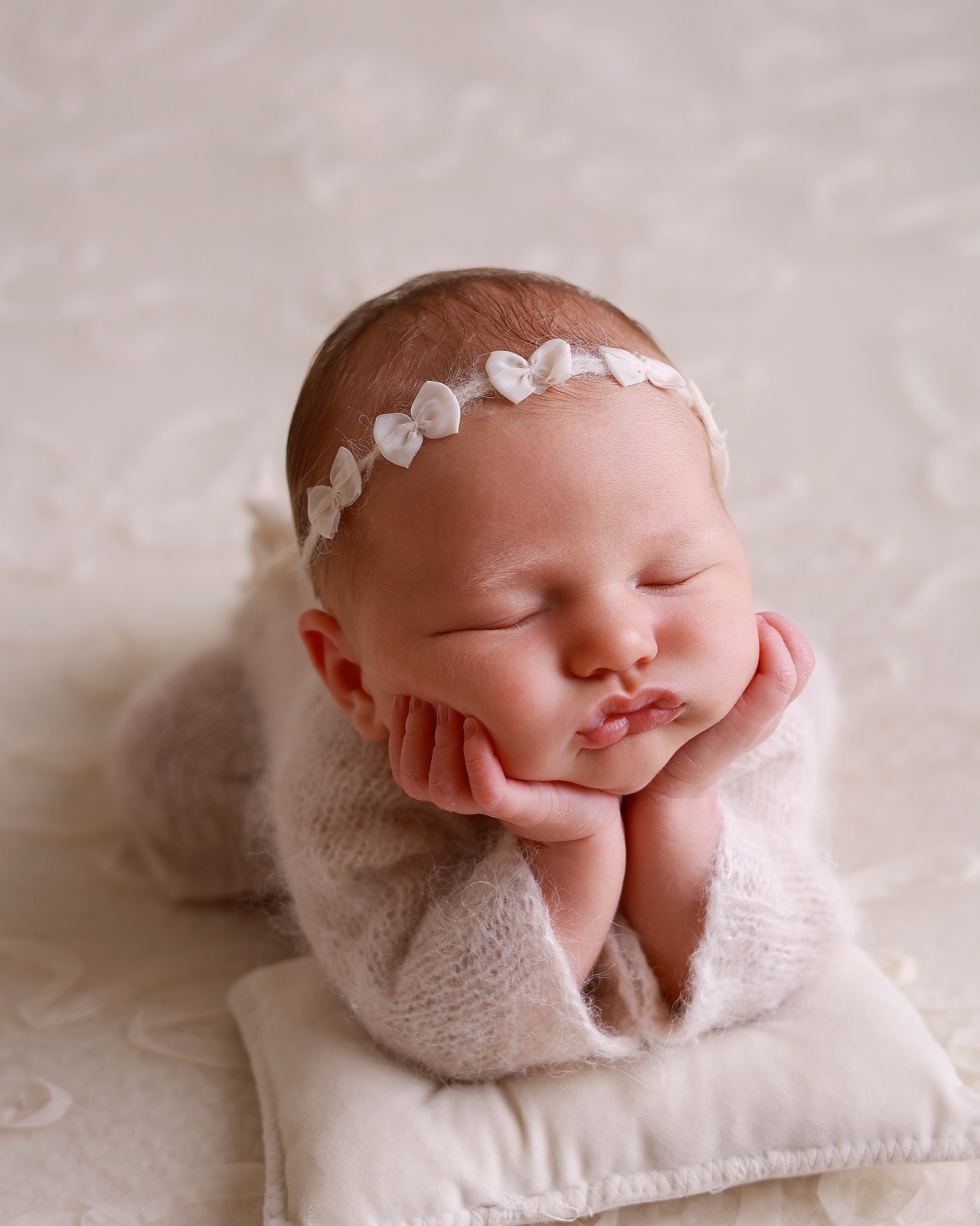 Close-up of a sleeping newborn resting her chin on her hands, tiny bow headband, cream cushion