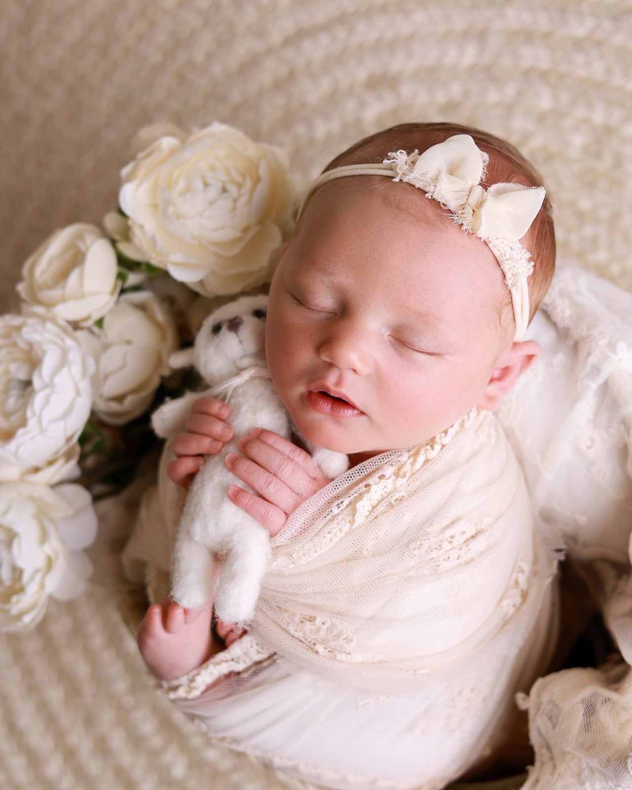 Sleeping newborn swaddled in cream lace, holding a small knitted teddy bear, surrounded by cream hydrangeas