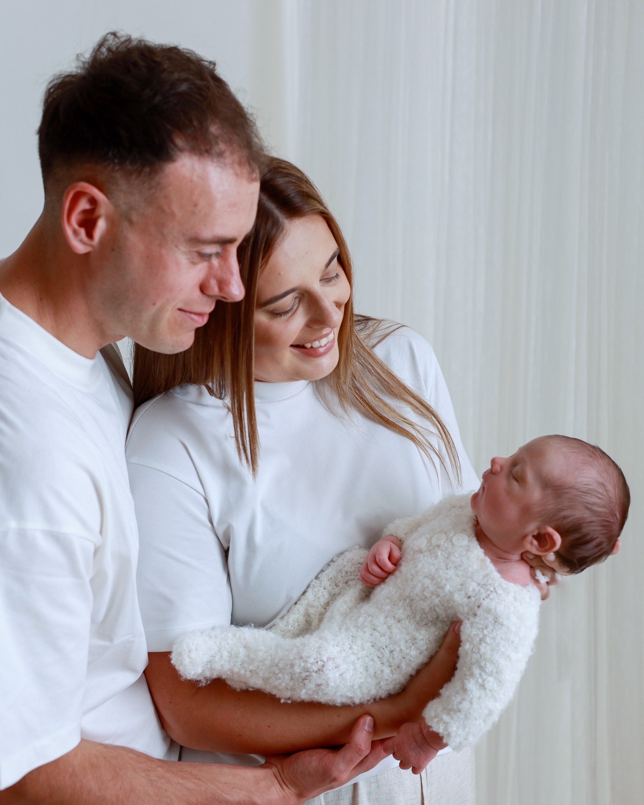 Mum and dad both smiling down at their sleeping newborn, cream knitted outfit, bright curtain backdrop
