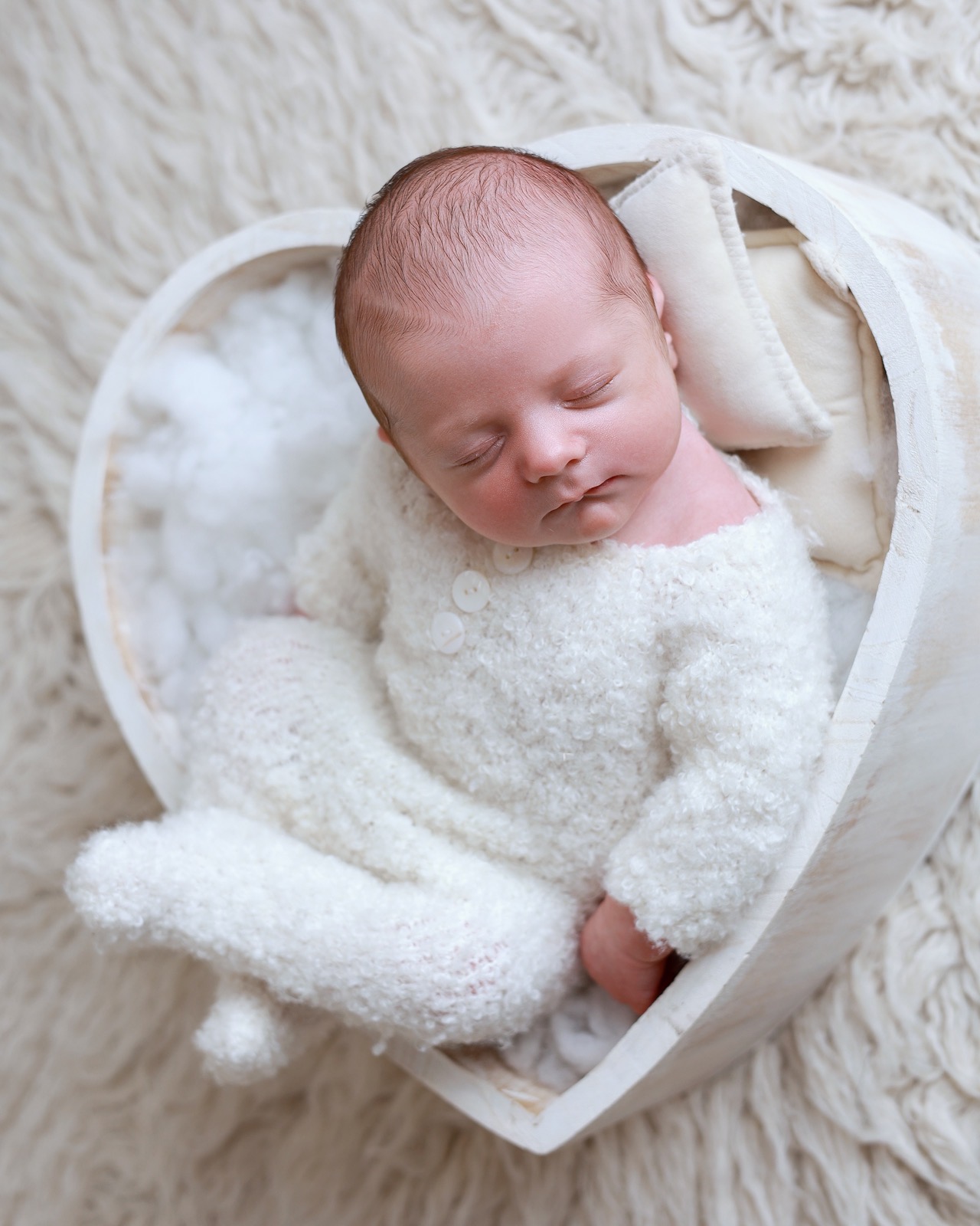 Sleeping newborn lying in a heart-shaped wooden prop, cream knitted outfit, cream faux fur backdrop
