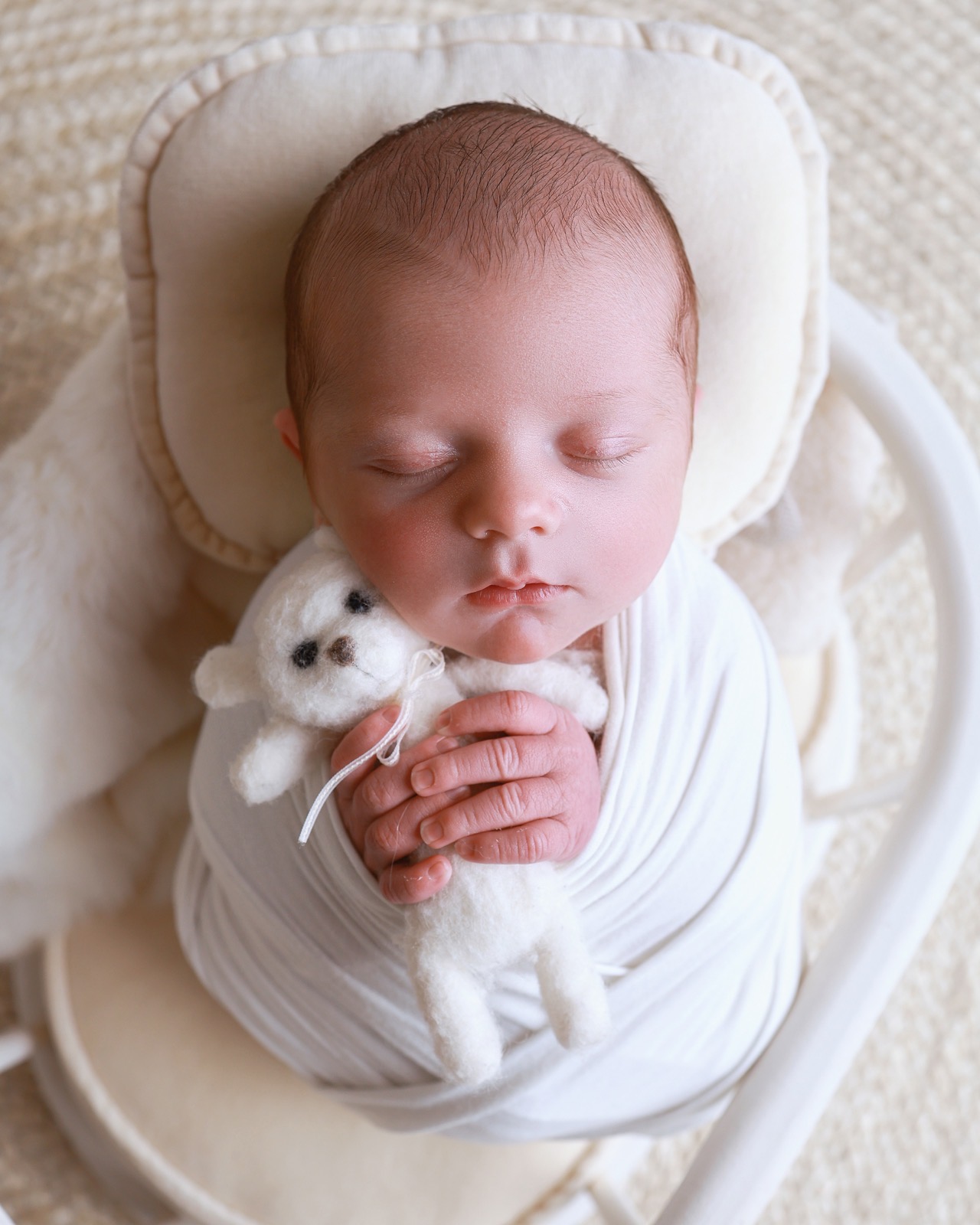 Sleeping newborn swaddled in a white wrap in a curved white basket, holding a small knitted teddy bear