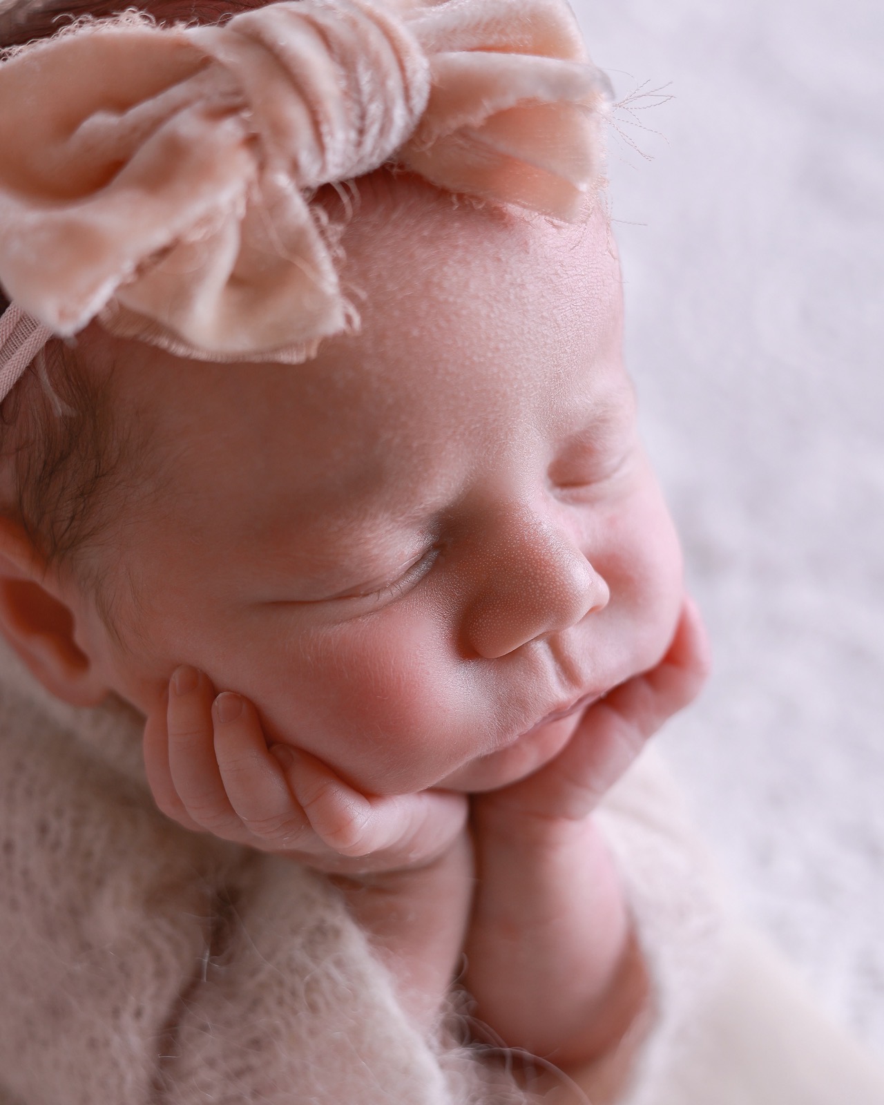 Extreme close-up of a sleeping newborn girl's face, peach velvet bow headband, chin resting on her hands