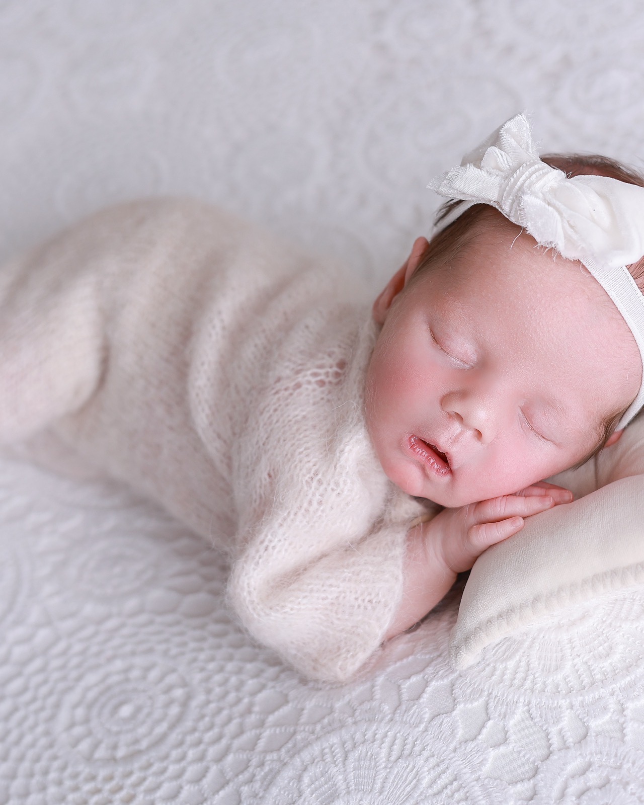 Sleeping newborn in side profile, cream bow headband, hand near her cheek, lace-textured backdrop