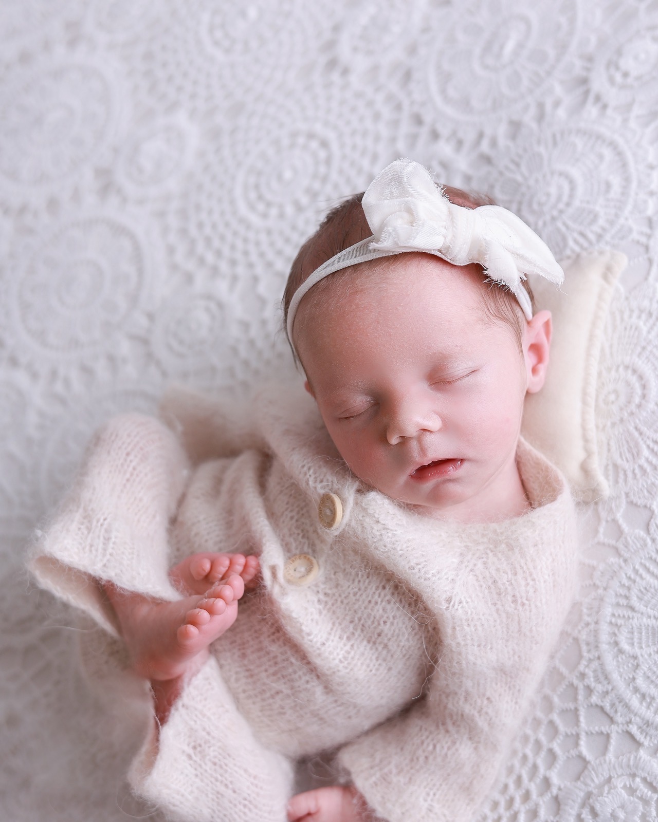 Sleeping newborn girl with one foot raised near her buttoned outfit, cream bow headband, lace-textured backdrop