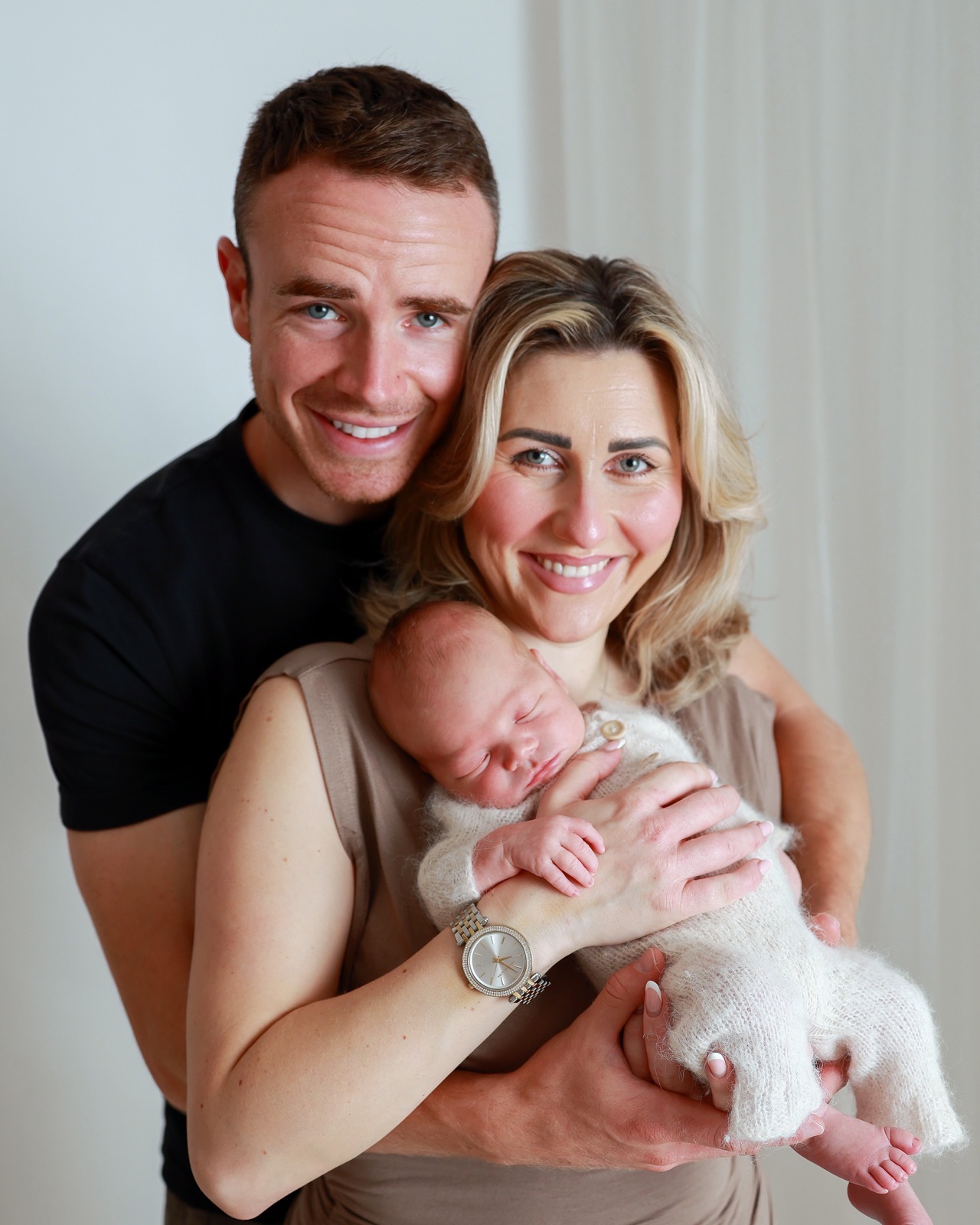 Mum and dad smiling down at their sleeping newborn, dad in a black t-shirt, bright curtain backdrop