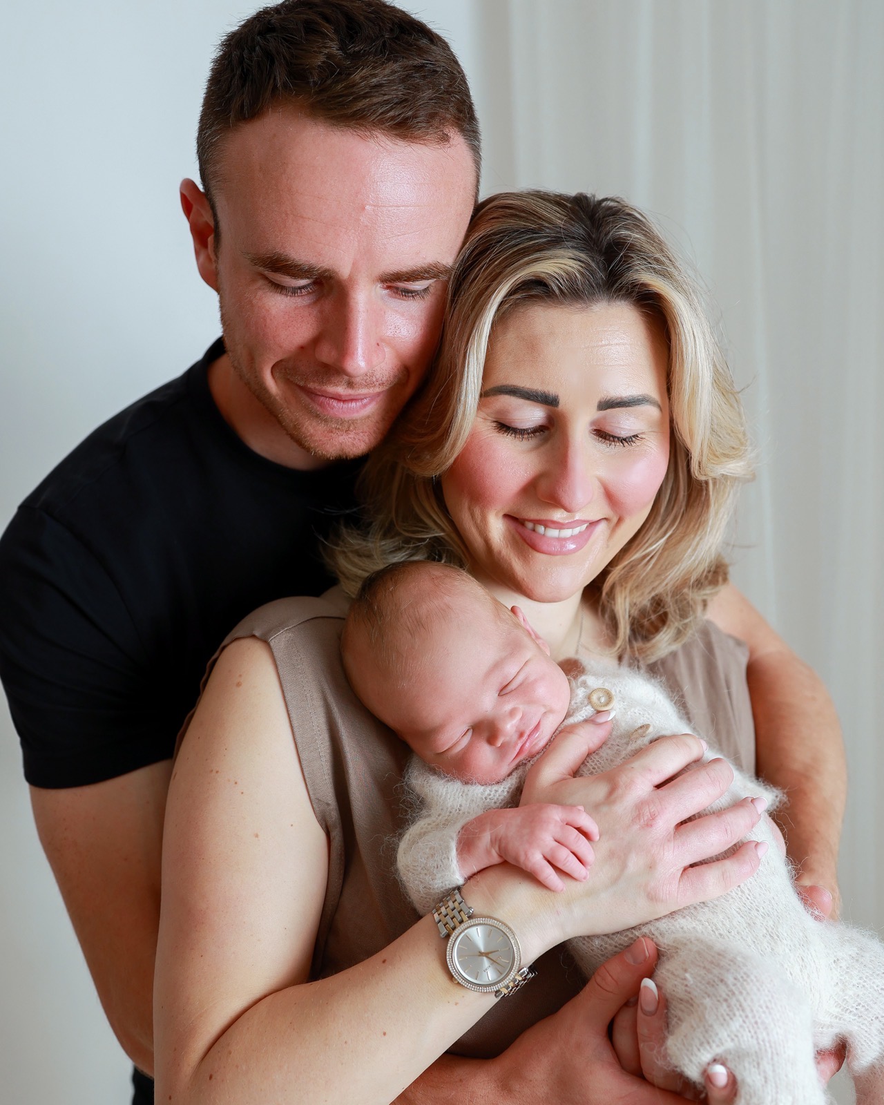 Mum and dad smiling closely at their sleeping newborn, mum's eyes closed, bright curtain backdrop