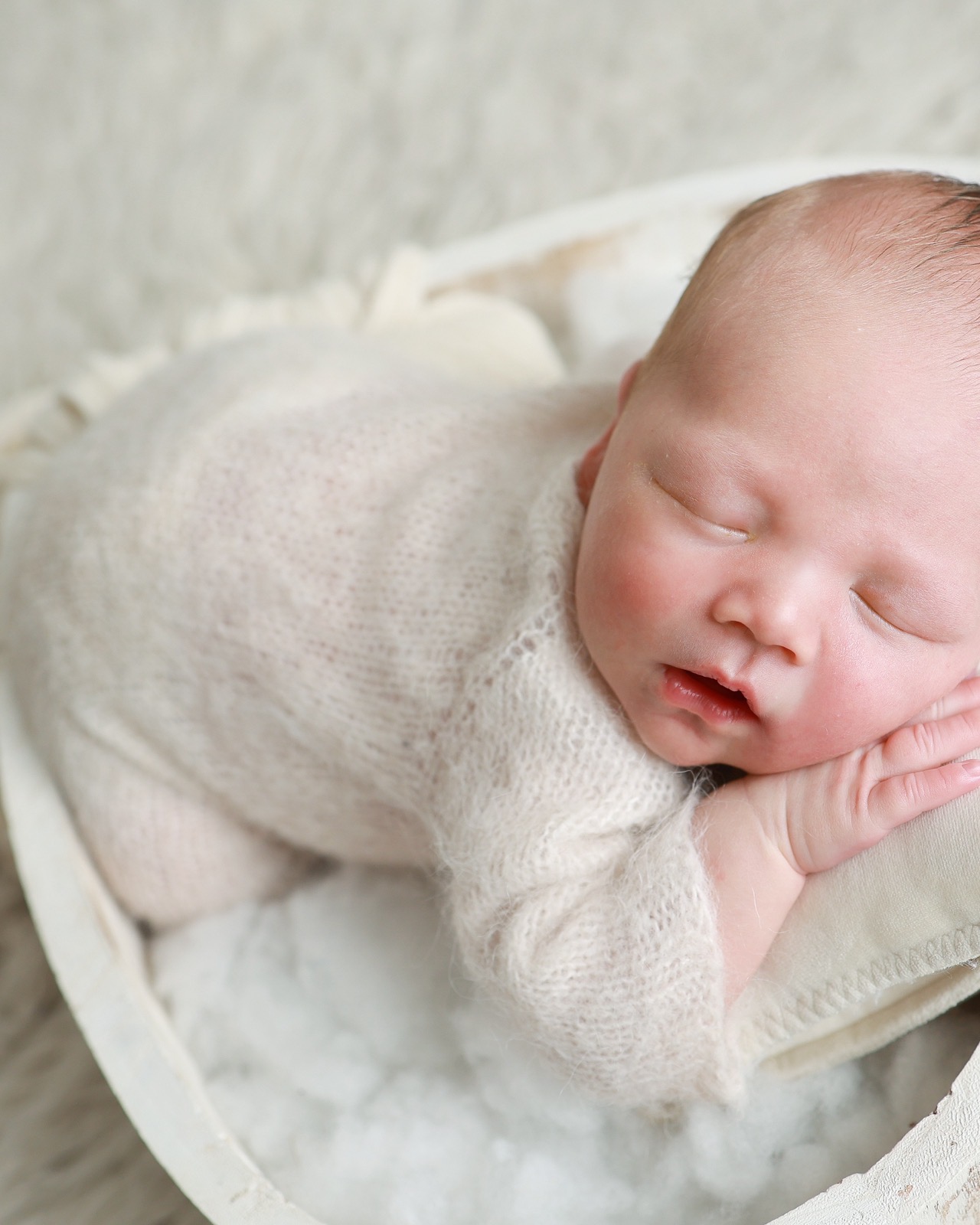Sleeping newborn boy in an oval wooden bowl, cream knitted outfit, hand resting near his cheek