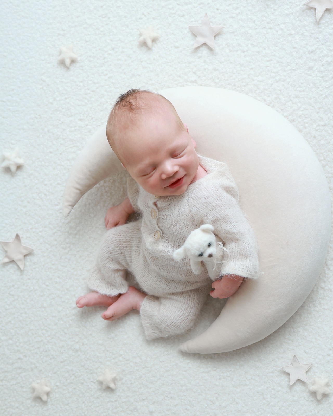 Newborn boy smiling in his sleep, lying in a crescent-moon prop, holding a small knitted teddy bear, star cut-outs nearby