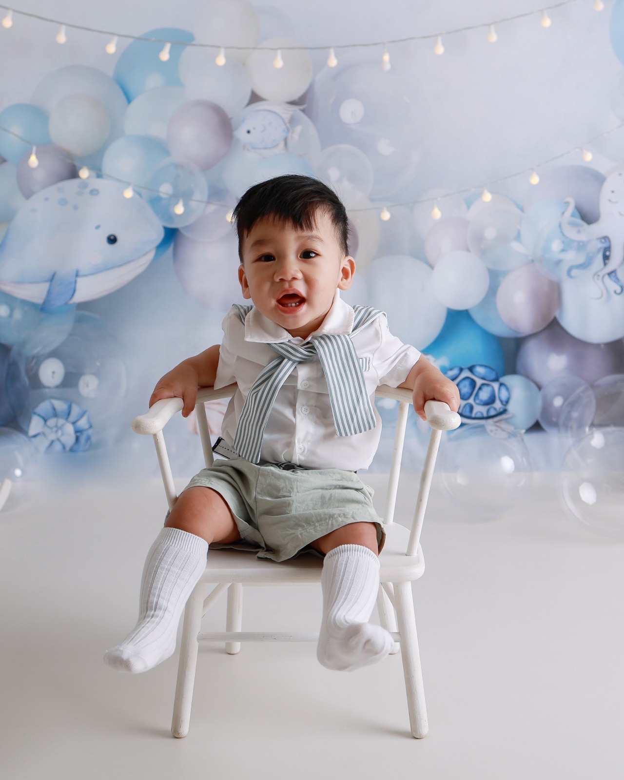 Toddler boy sitting beside a small birthday cake, wearing a white sailor-style bow tie, ocean-themed balloon backdrop