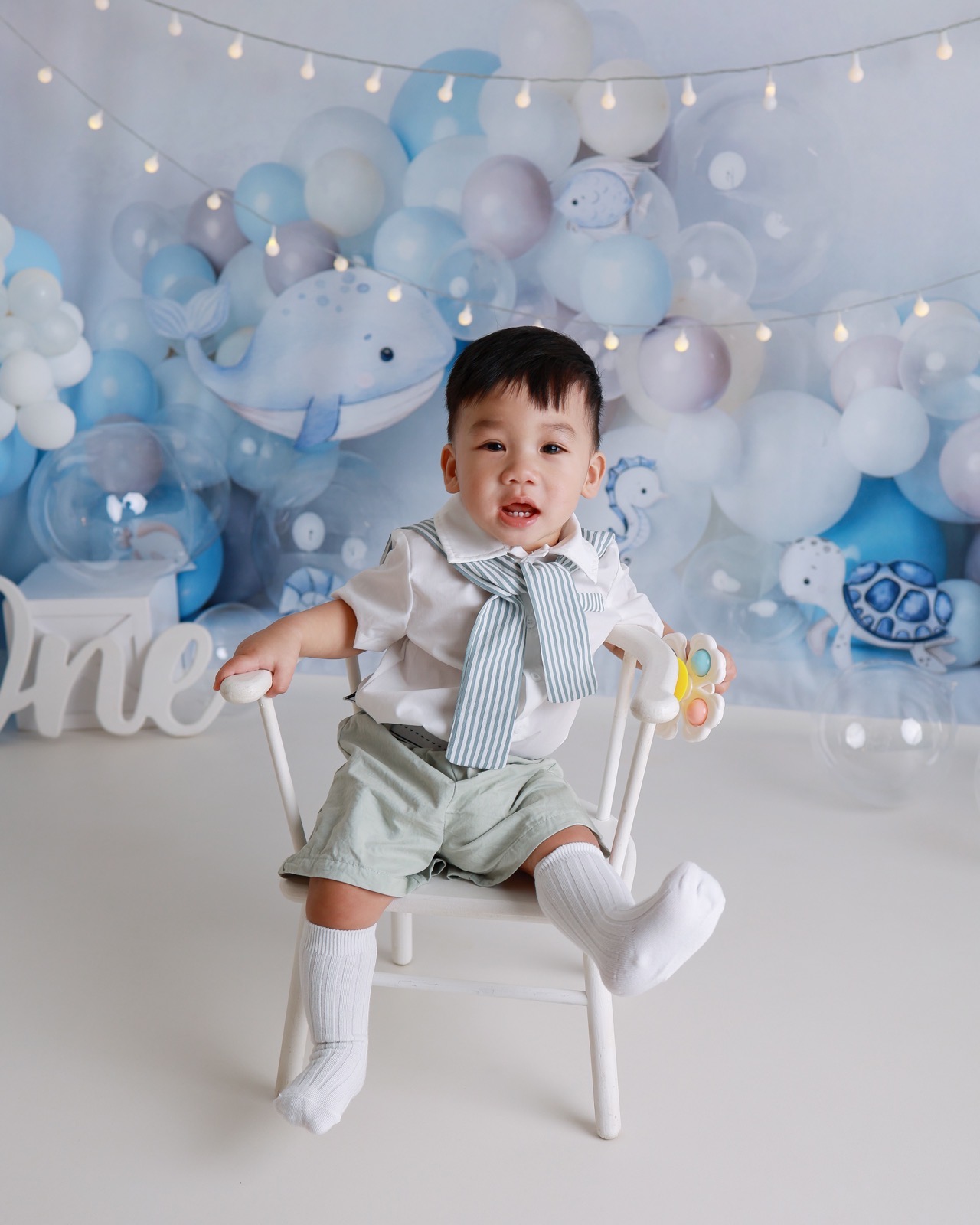 Toddler boy in a sailor-style bow tie holding a rattle, sitting on a white chair, ocean-themed balloon backdrop