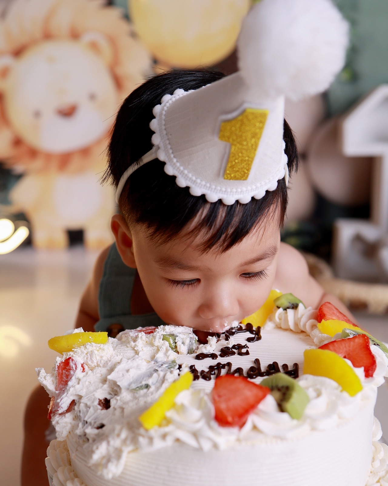 Close-up of a toddler boy with his face in a cake decorated with fresh fruit, '1st Birthday' piped in chocolate