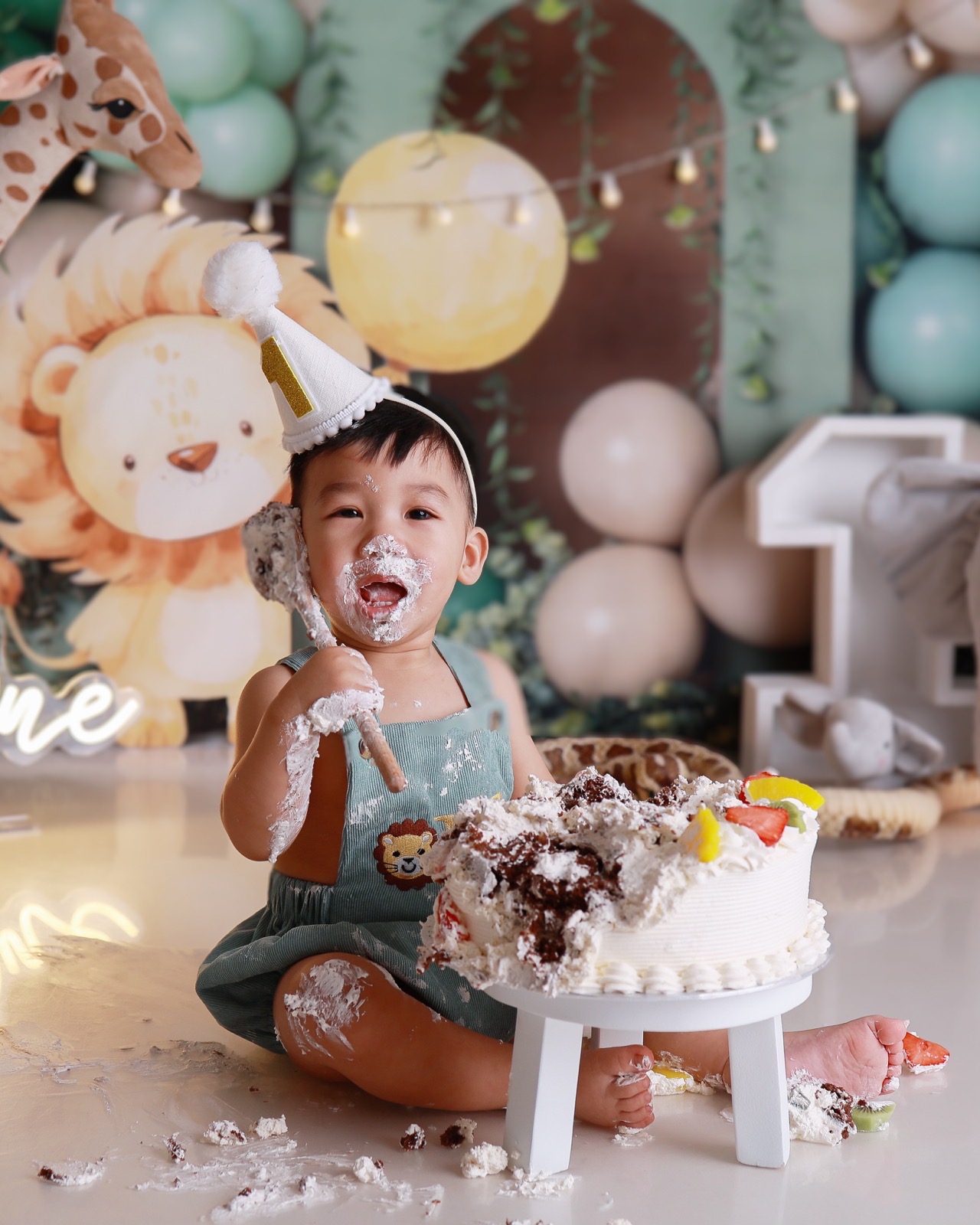 Toddler boy covered in cake with a wooden spoon, cake mostly demolished, safari-themed balloon backdrop