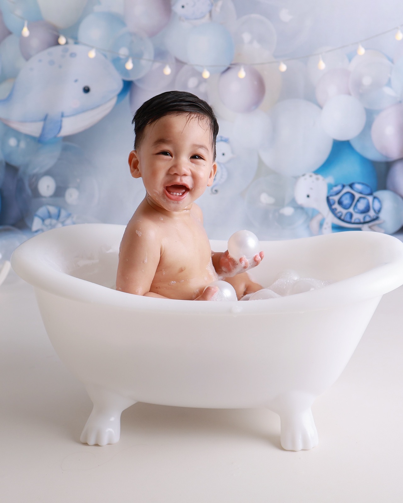 Toddler boy laughing in a white clawfoot bath tub full of bubbles, ocean-themed balloon backdrop